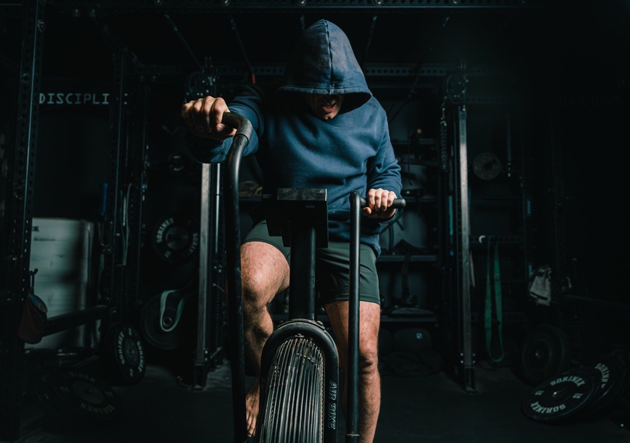 A person wearing a hoodie and shorts, with their face partially hidden under the hoodie, riding a stationary airbike in a dark gym.