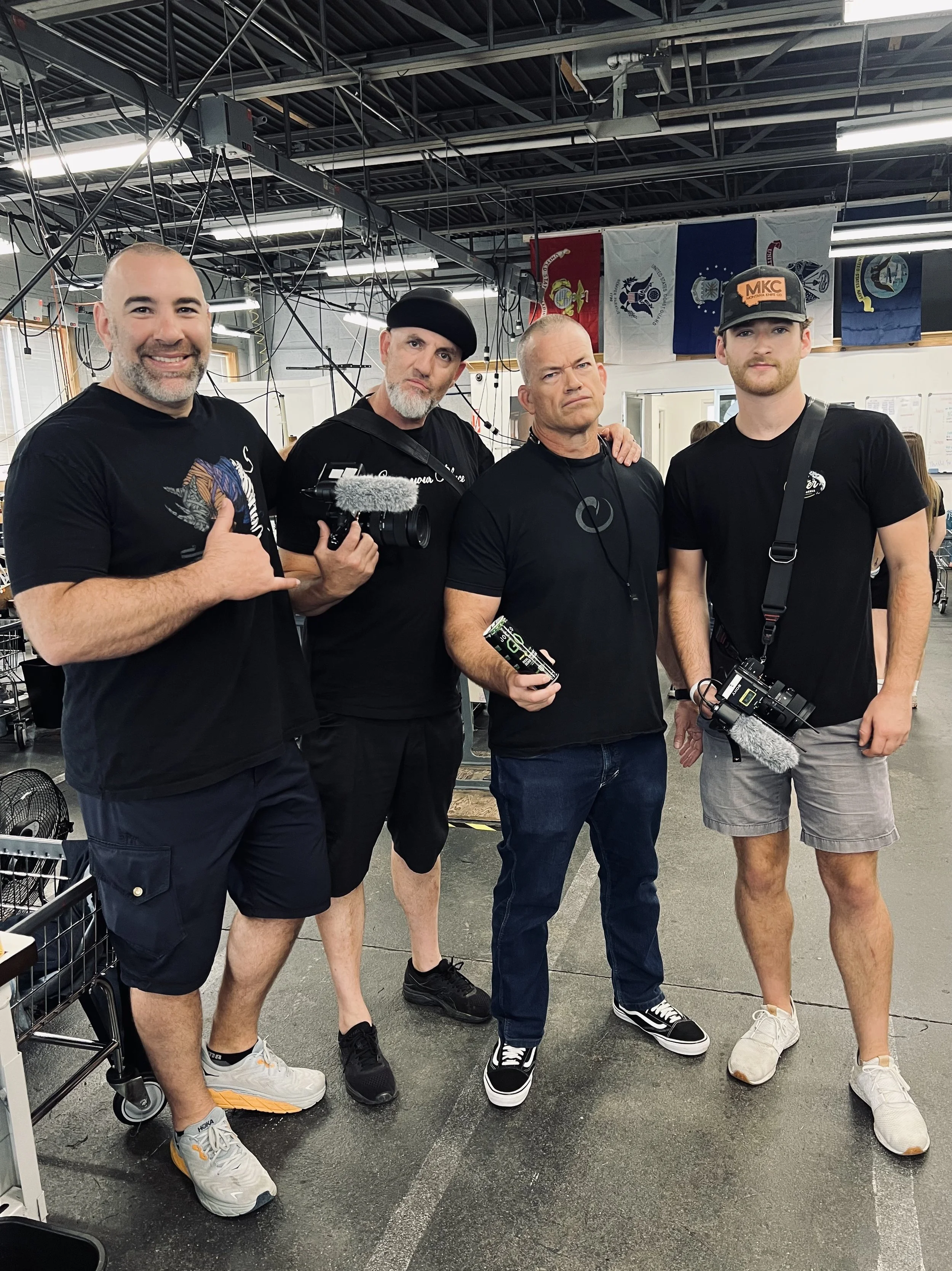 Four men standing together in a room with flags hanging in the background. They are dressed casually, some holding cameras and equipment, suggesting they are part of a media or filming crew.