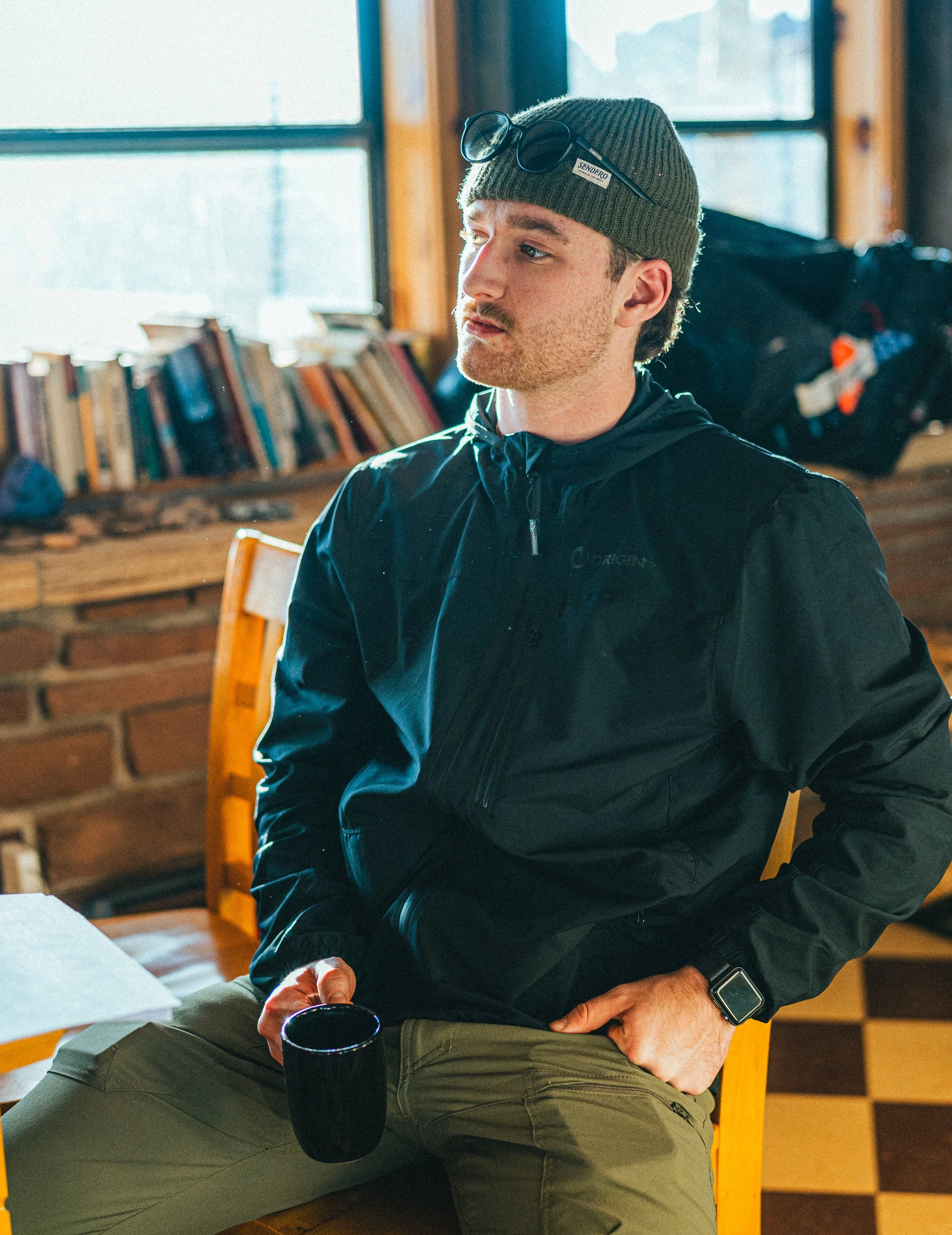 Young man sitting in a cozy, rustic room holding a black mug, wearing a green beanie, sunglasses, black jacket, and tan pants, with a bookshelf and window in the background.