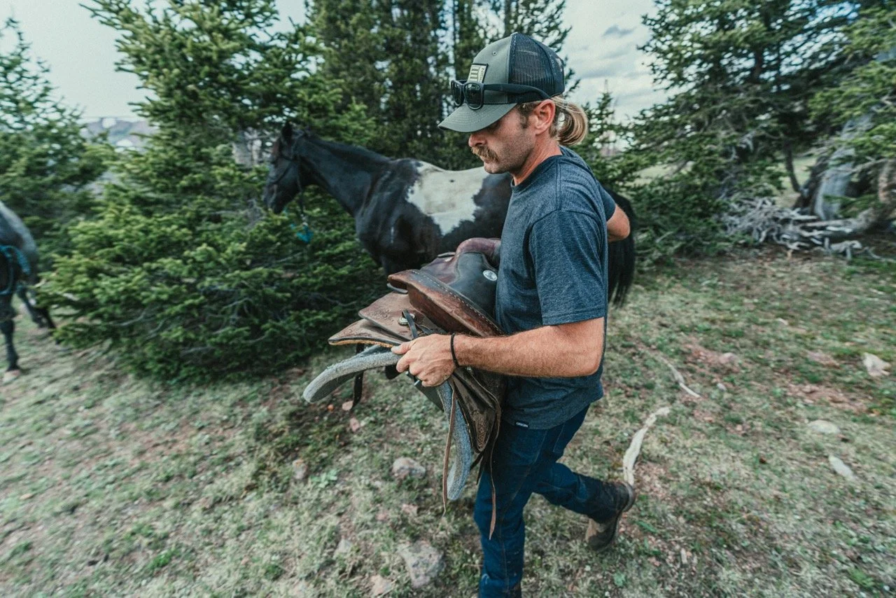A man outdoors in a wooded area carrying a saddle, with a black and white horse in the background.