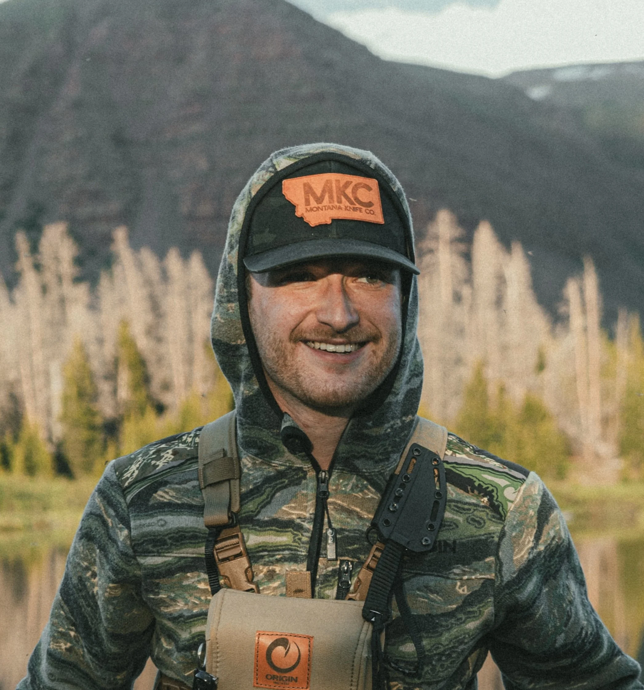 A man outdoors in a camouflage jacket, wearing a black cap with an orange patch, and smiling, with mountains, trees, and a lake in the background.
