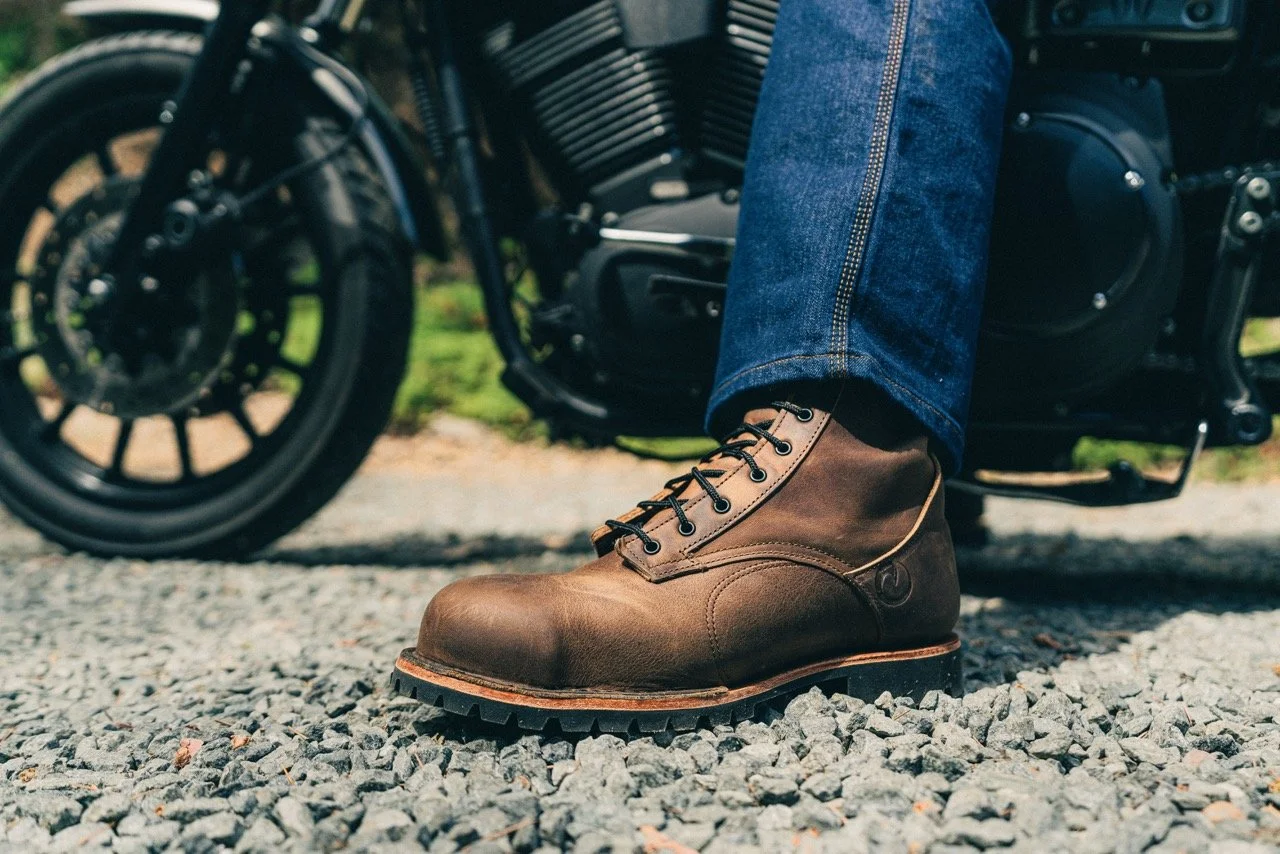 A person wearing brown leather work boots and blue jeans, standing on gravel ground in front of a black motorcycle.