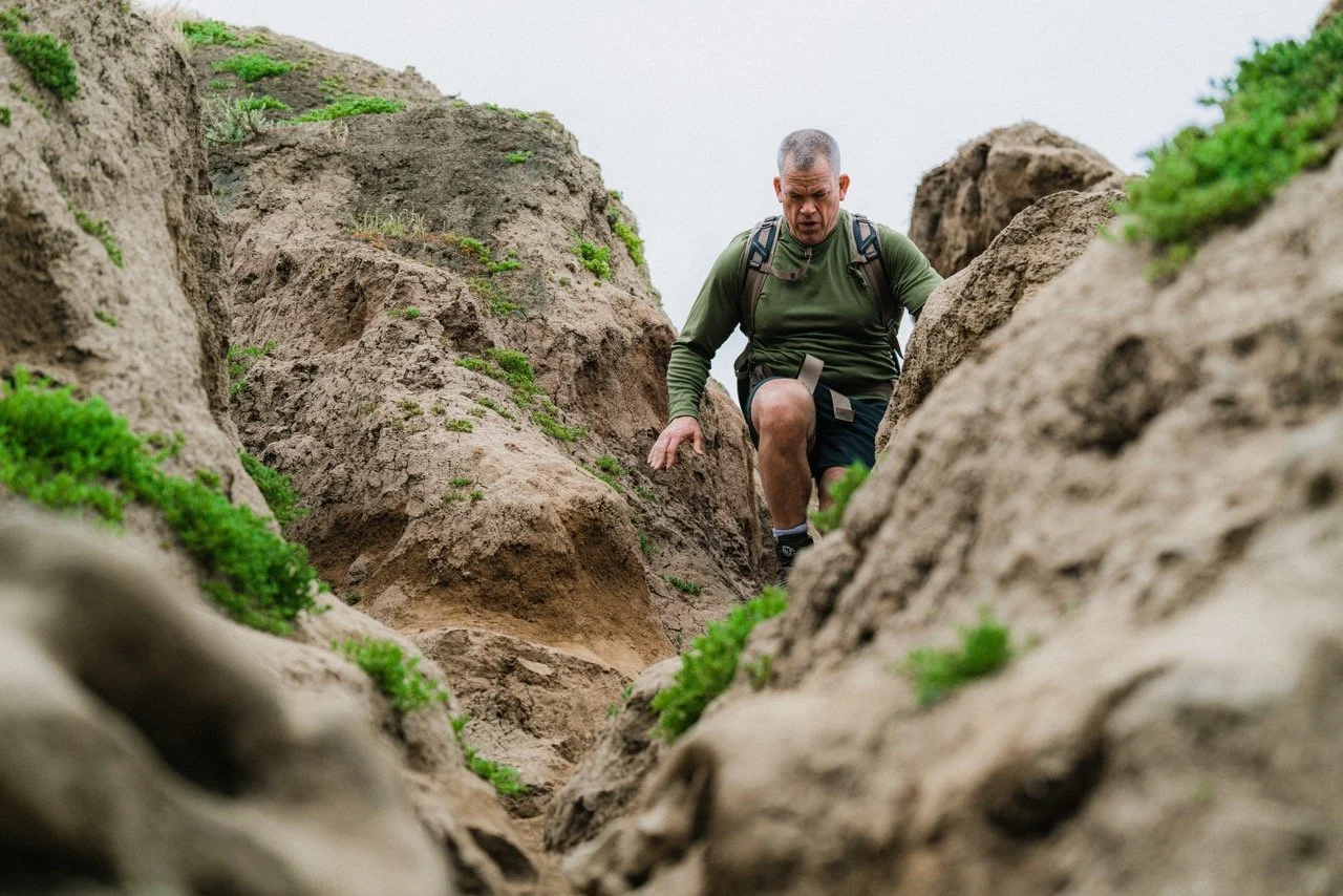 Man hiking up a rocky trail with green plants and gray sky.