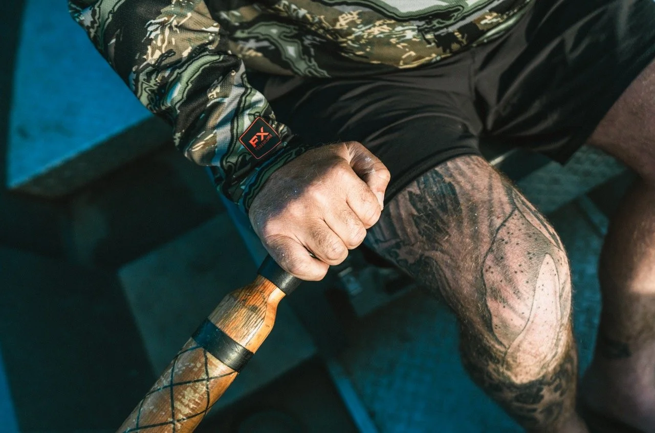 A person with a camouflage jacket holding a wooden stick, wearing dark camouflage shorts, sitting on a blue surface.