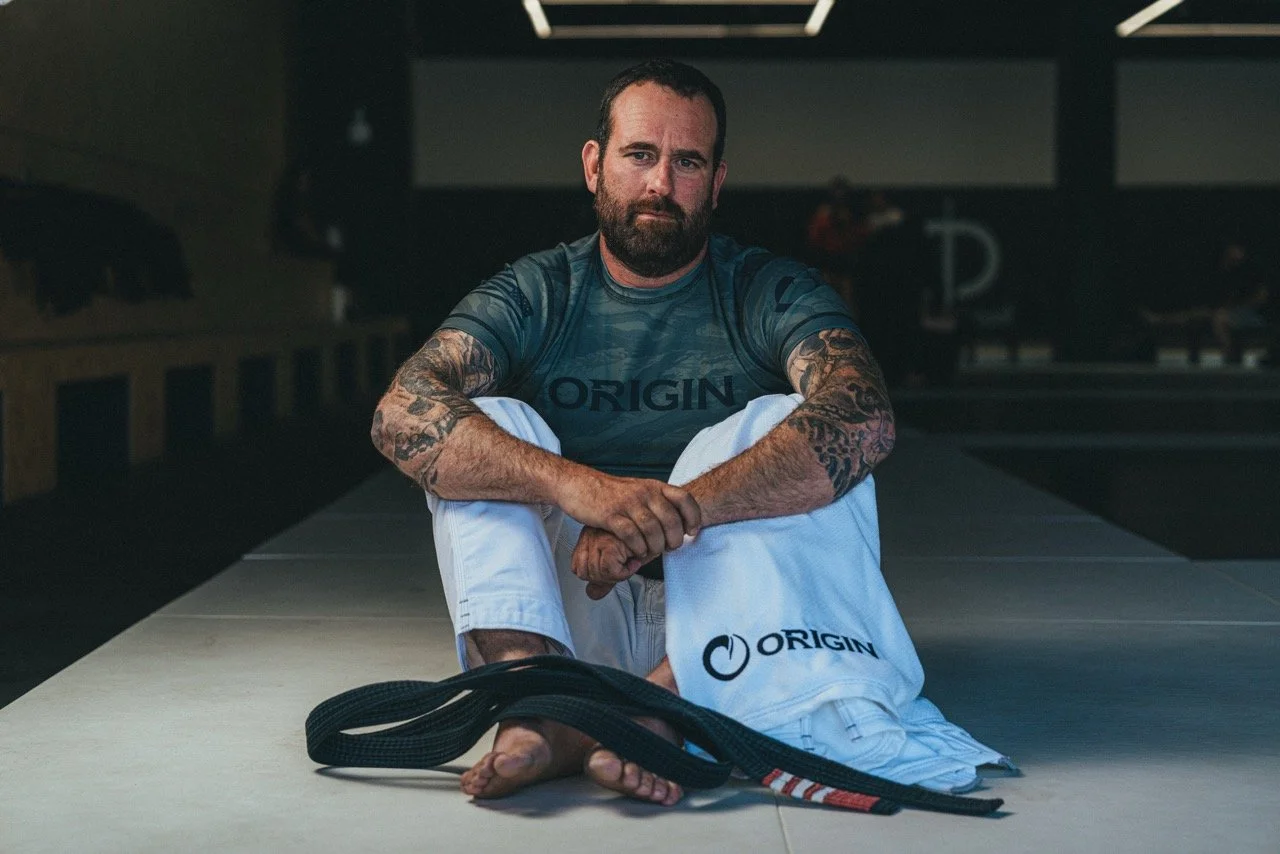 A man with tattoos sitting on a martial arts mat, wearing a blue shirt with 'ORIGIN' logo and white martial arts pants with the same logo, in a gym or dojo setting with a black belt placed in front of him.