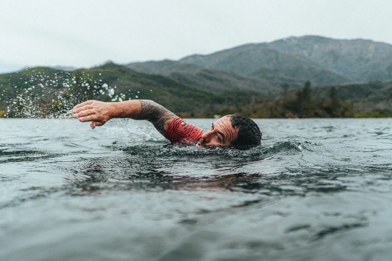 A man swimming and doing the freestyle stroke in an outdoor natural body of water with mountains in the background.