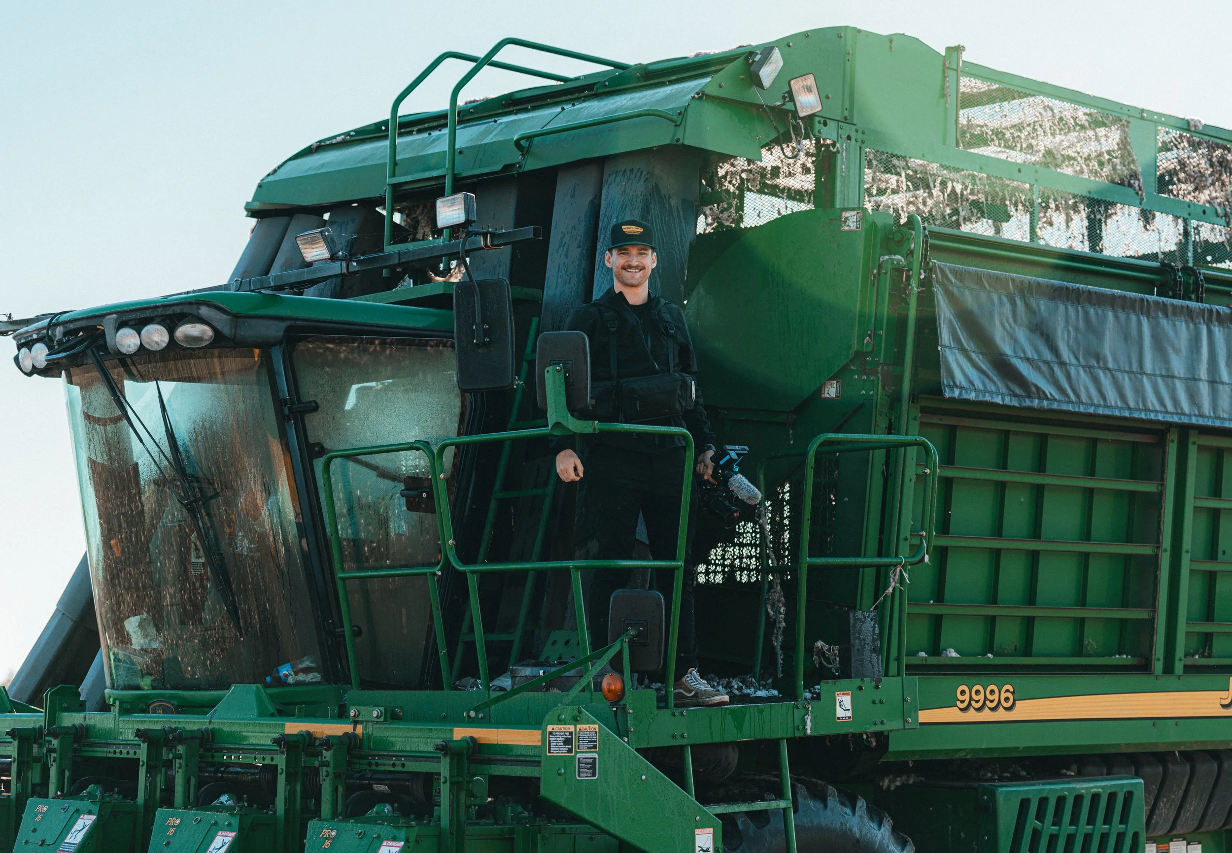 Person standing on a green John Deere combine harvester, smiling, wearing black clothing and a hat.