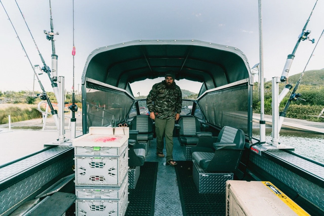 A man in camouflage jacket and green pants standing inside a covered fishing boat with multiple fishing rods mounted on the sides, water and greenery visible in the background.