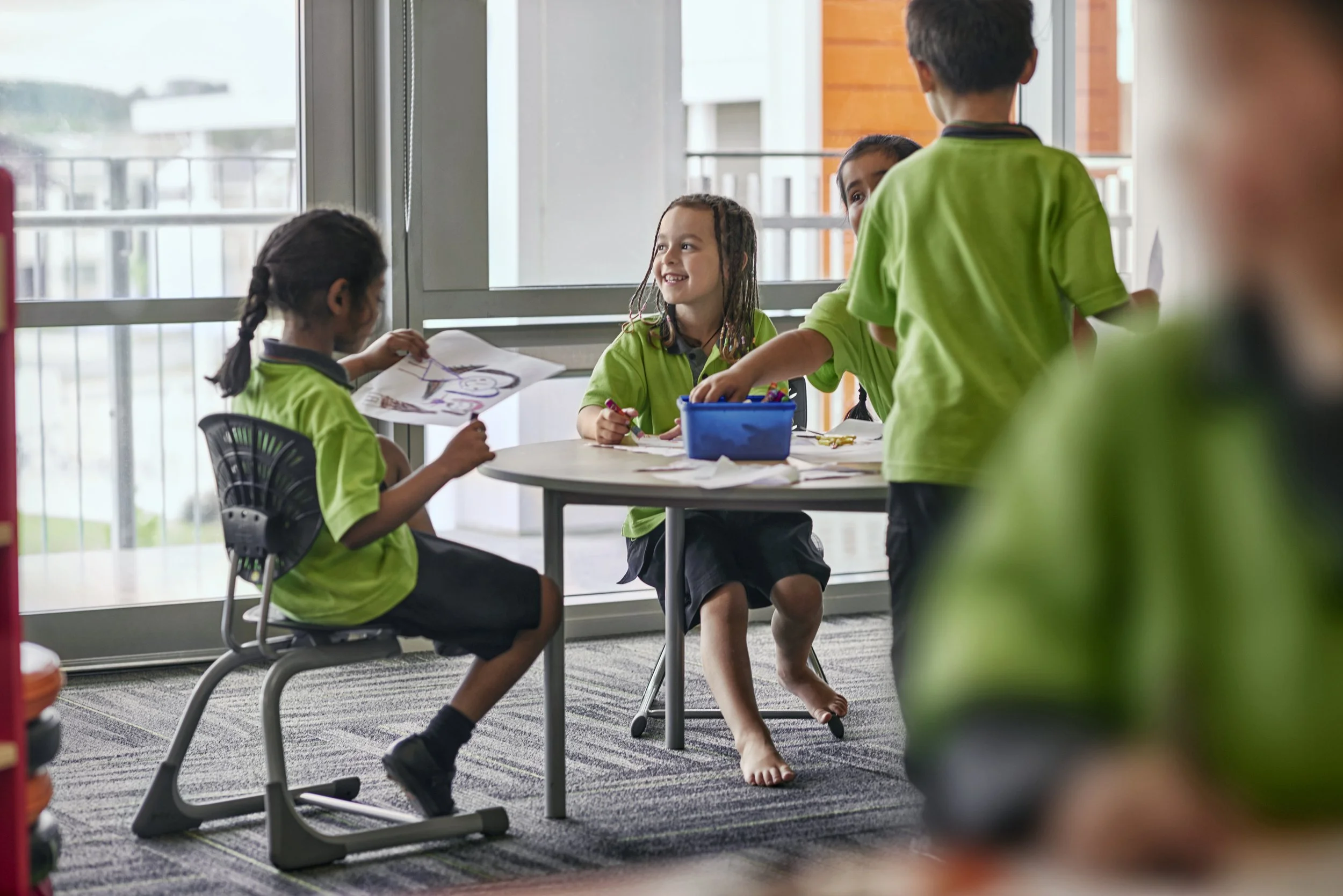 Group of young children in green uniforms engaging in arts and crafts at a table in a classroom with large windows.
