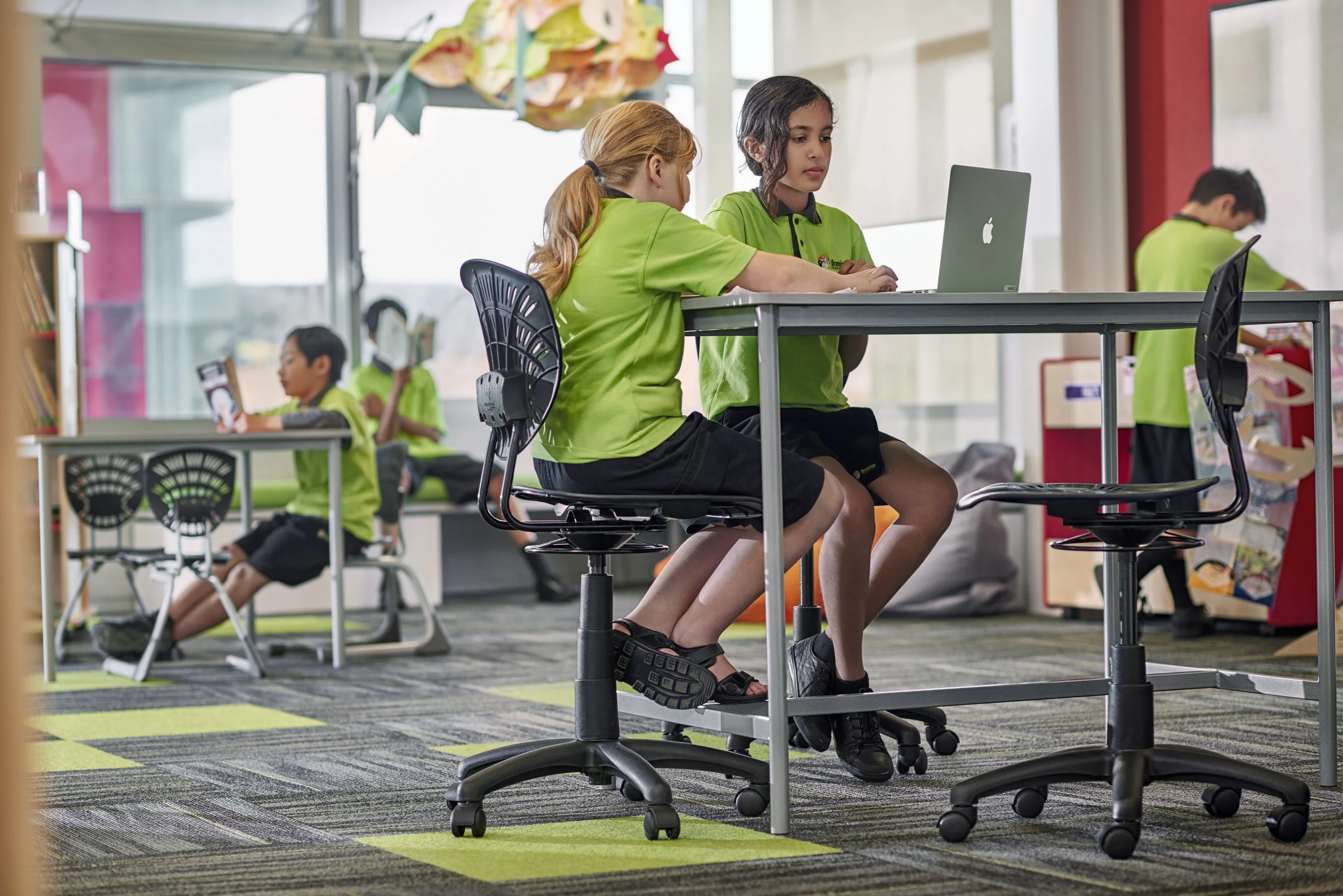 Children in school uniforms working on laptops and tablets in a classroom with large windows and colorful decorations.