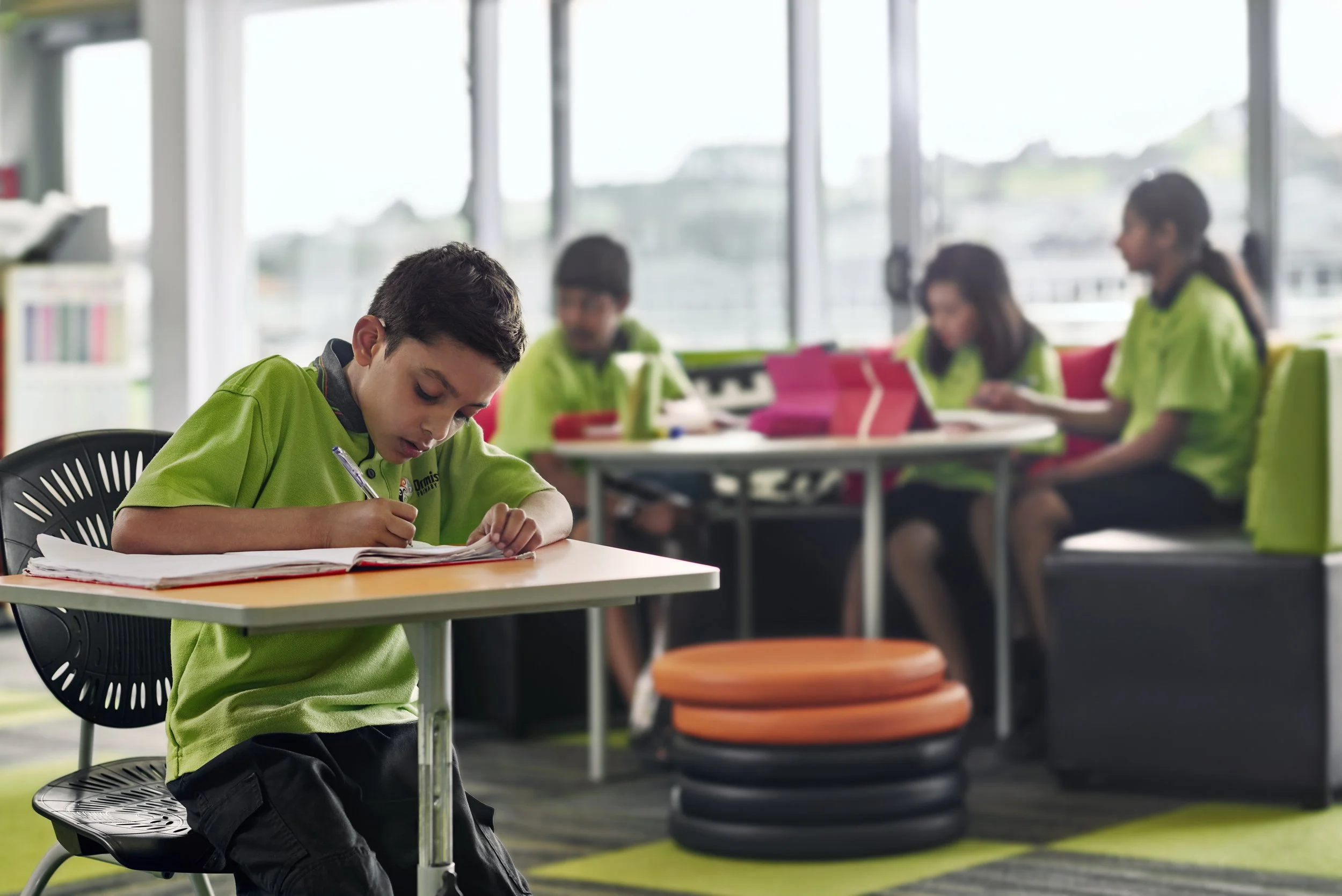 Young boy in green school uniform writing in a notebook at a table inside a classroom with large windows, with other students working in the background.