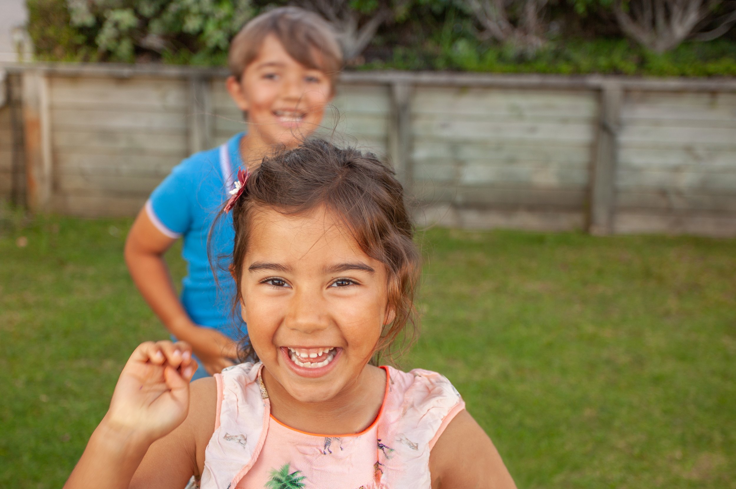 Two children in a backyard, smiling and playing. The girl in front has brown hair with a pink hair clip, wearing a pink shirt with green palm trees. The boy behind, with light brown hair, is wearing a blue shirt. They are near a wooden fence and some