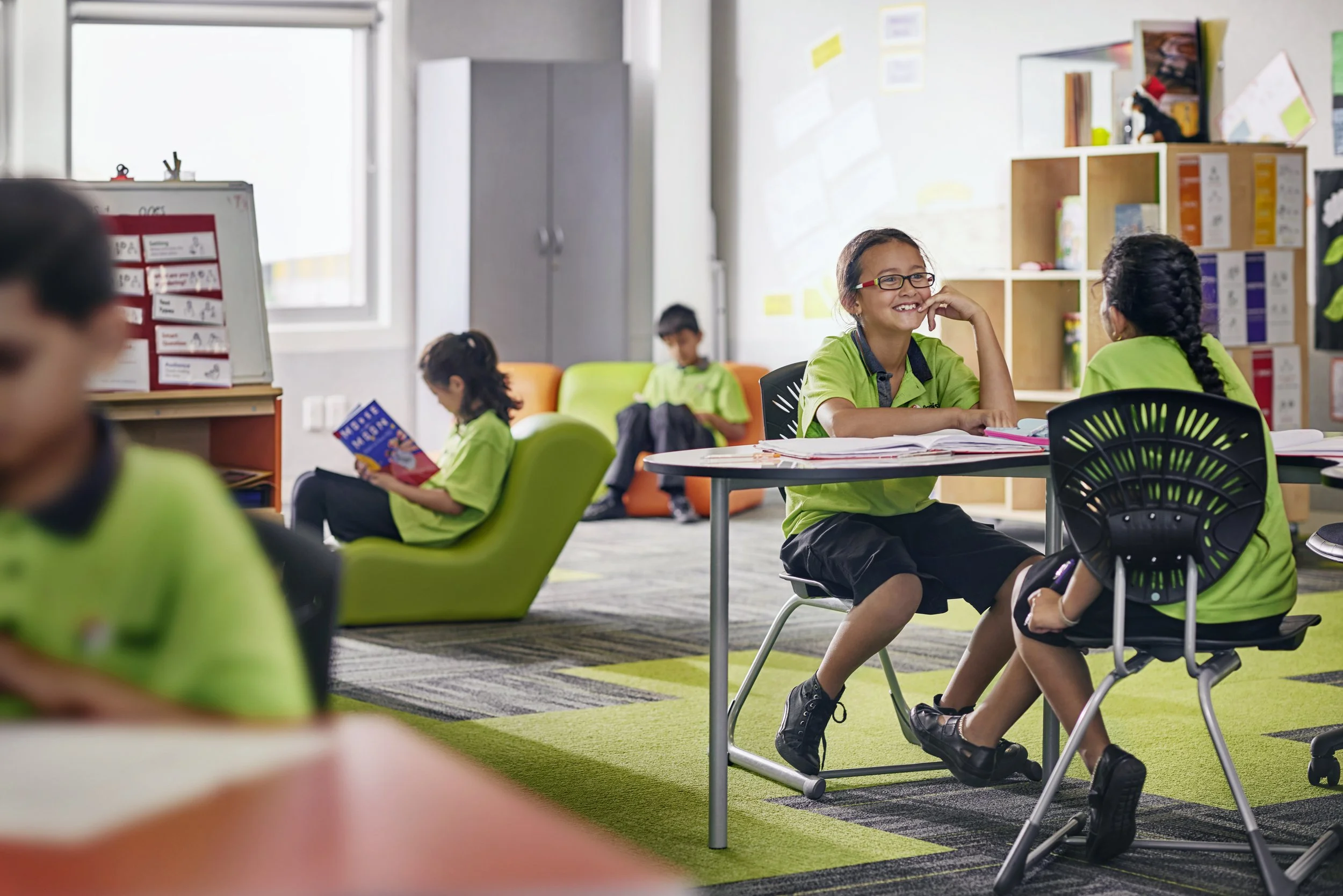 Smiling young girl with glasses sitting at a table in a classroom, talking to another girl. Other students are reading and studying in the background.