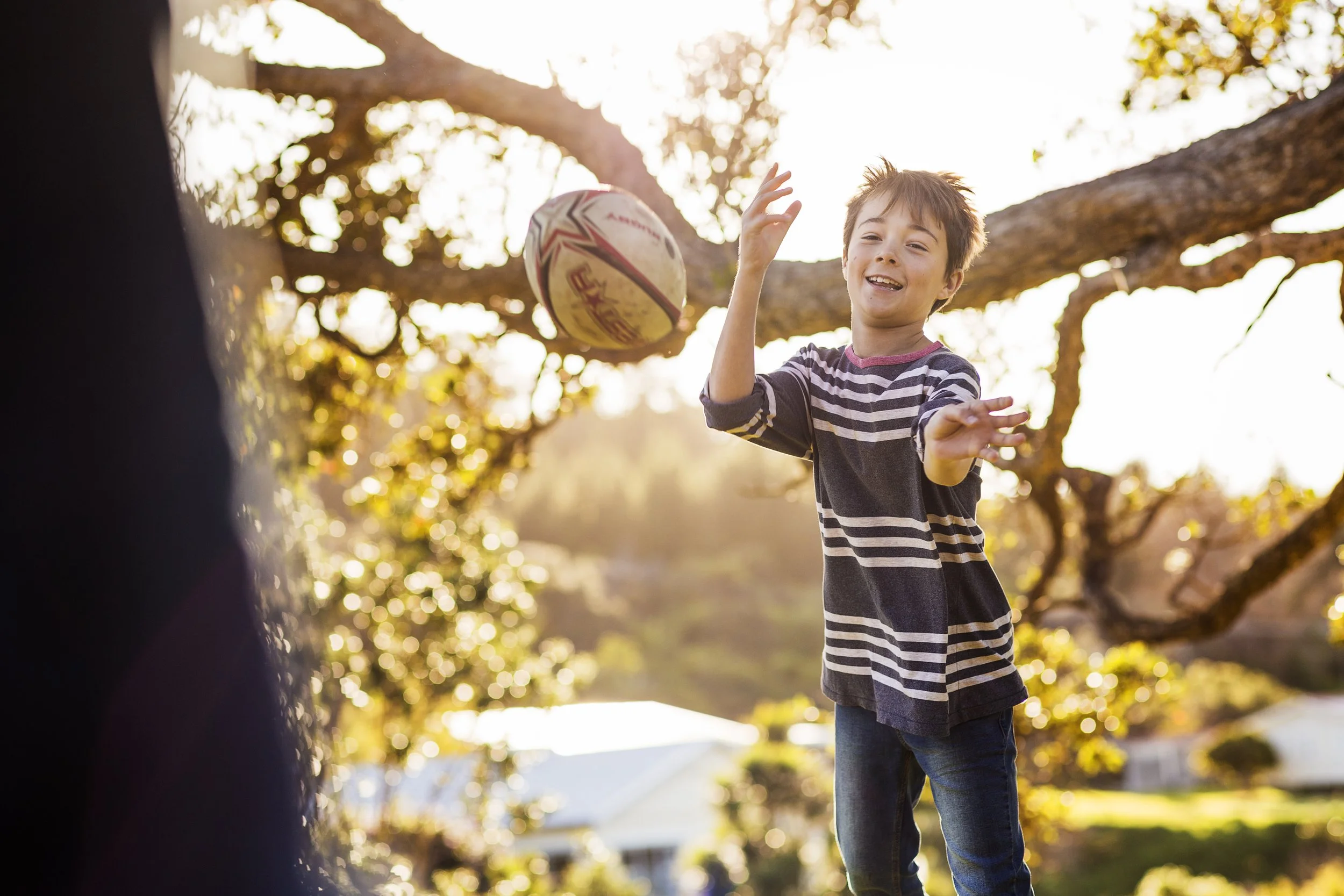 A boy playing with a rugby ball outdoors on a sunny day, with trees and houses in the background.