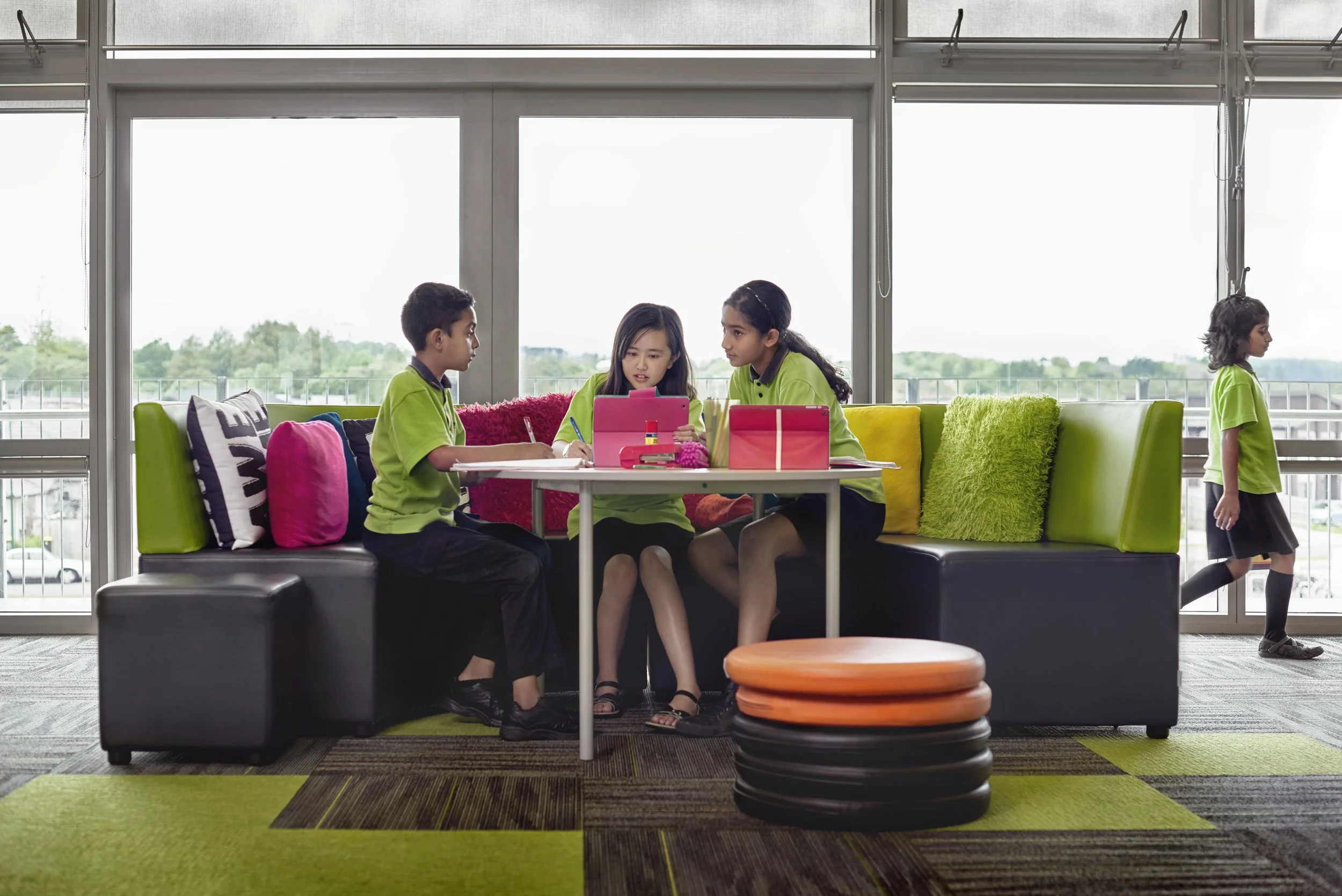 Four children sitting around a table inside a brightly lit room, with one girl showing something on a pink tablet to two other girls, and a boy sitting nearby. Another girl is walking past behind the sofa, which has colorful pillows, and there are bl