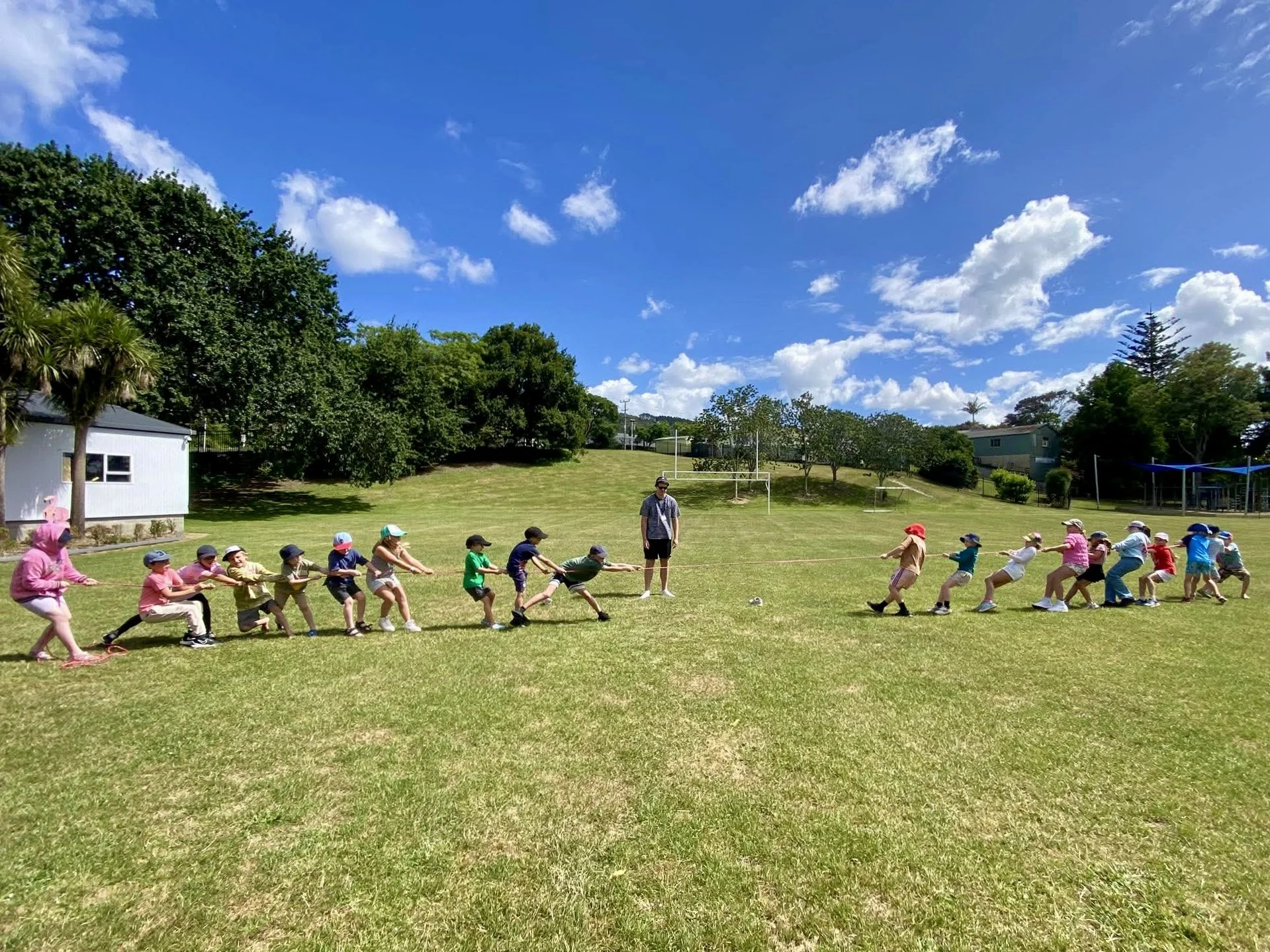 Children playing tug of war on a grassy field with a blue sky and scattered clouds, surrounded by trees and a few buildings in the background.