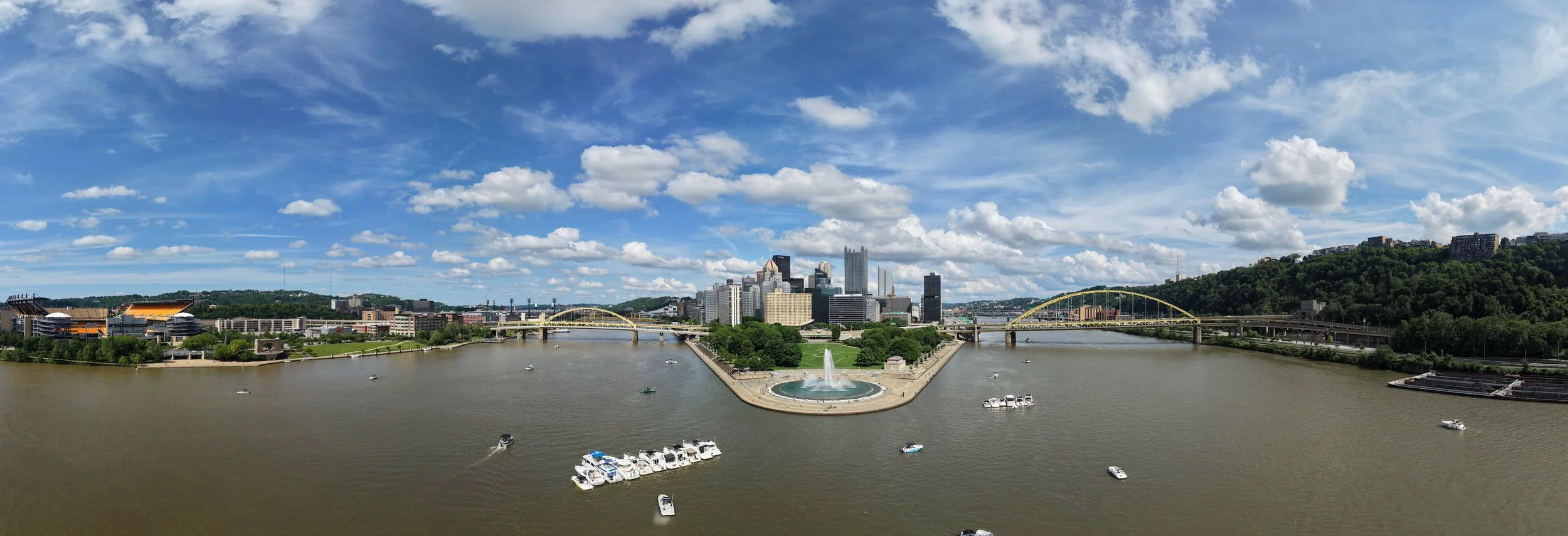 Panoramic view of Pittsburgh skyline across the Allegheny River on a partly cloudy day, with boats on the water, green hills, and bridges visible.