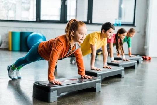 Four children exercising on aerobic steps in a gym.