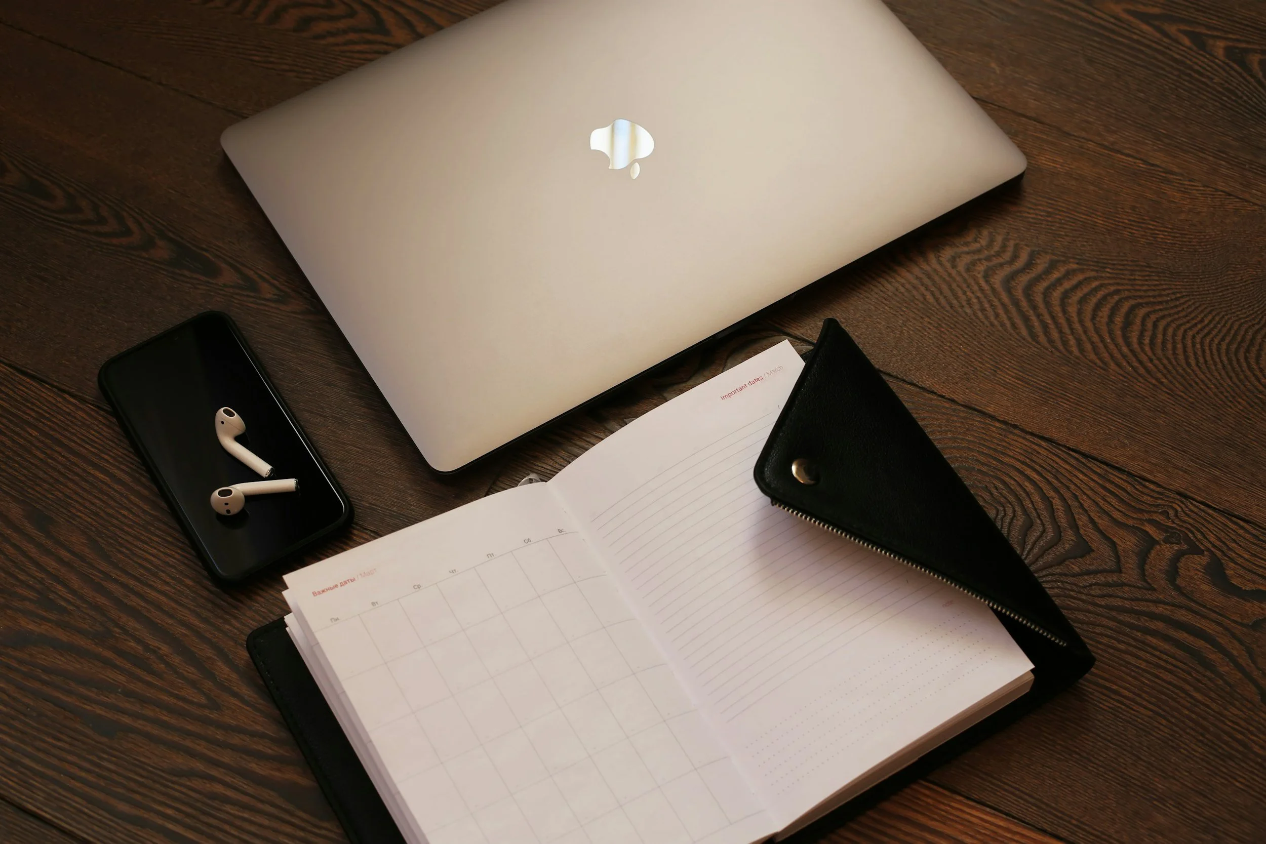 Open planner with lined and dotted pages, black leather cover, MacBook laptop with Apple logo, smartphone with wireless earbuds, black zippered wallet on a wooden desk.