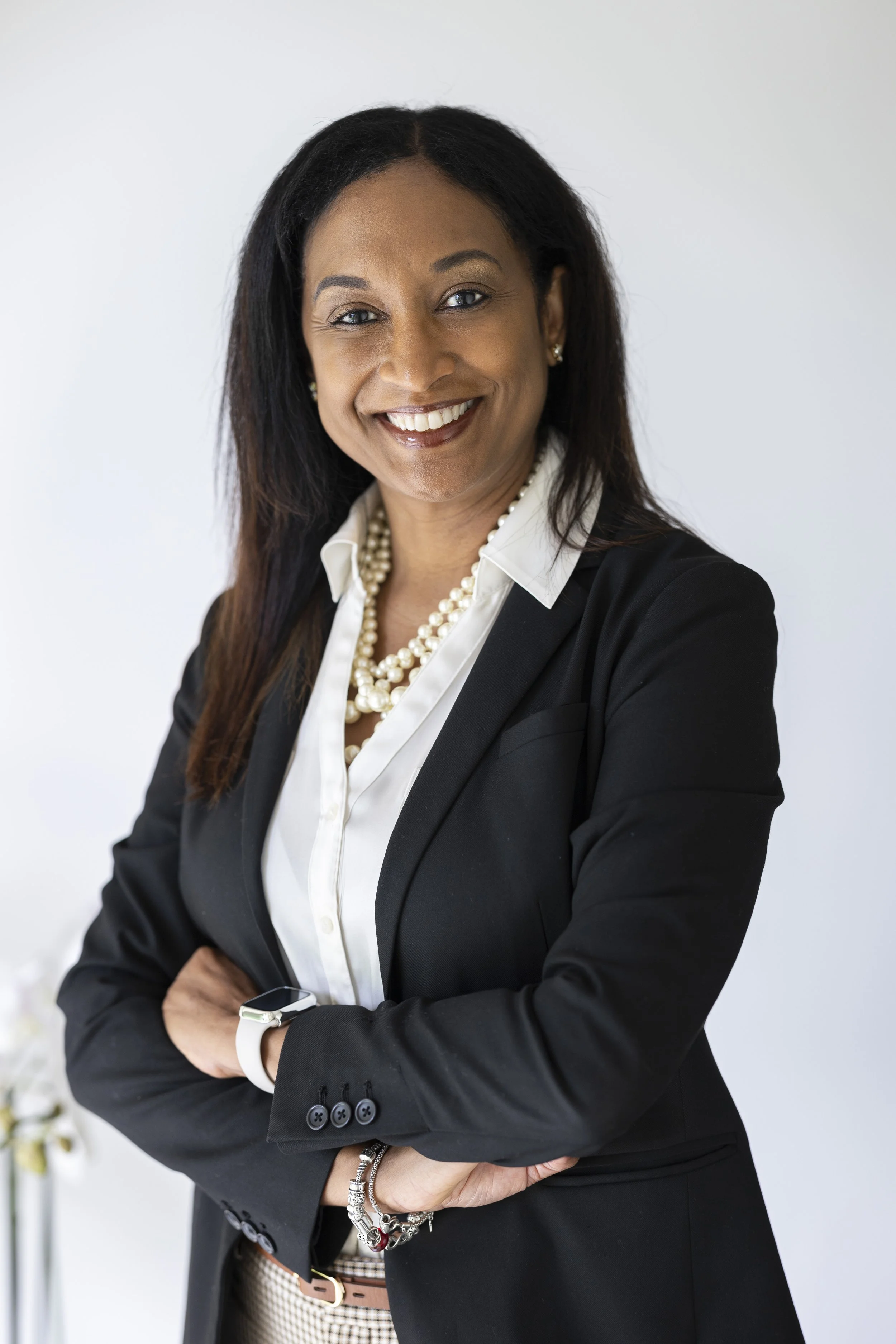 A professional woman with dark hair, wearing a black blazer, white blouse, and pearl necklace, smiling with arms crossed against a plain white background.