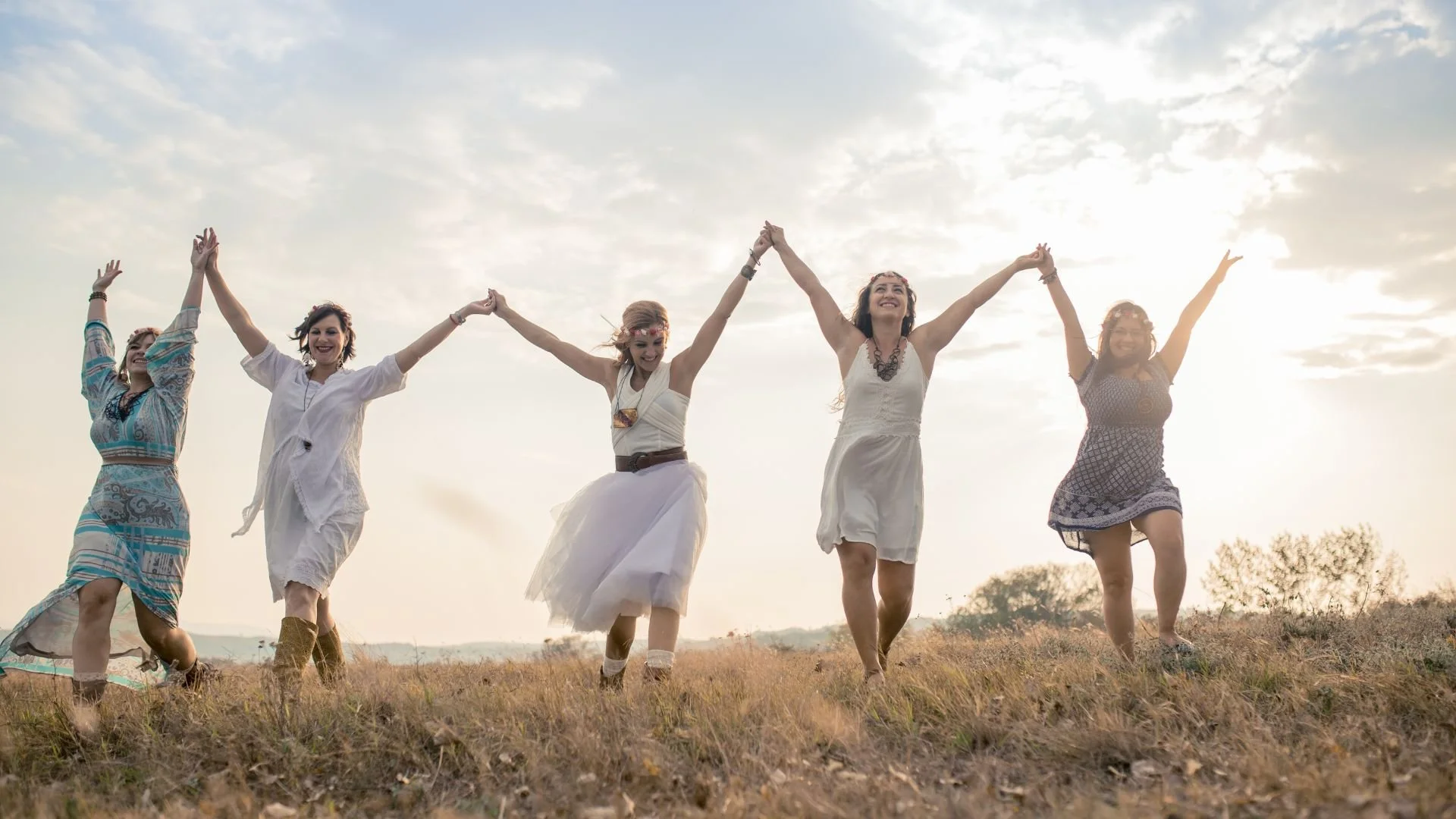 Five women holding hands and celebrating as they journey together, supporting each other achieving their goals, outdoors in a grassy field with a cloudy sky in the background.