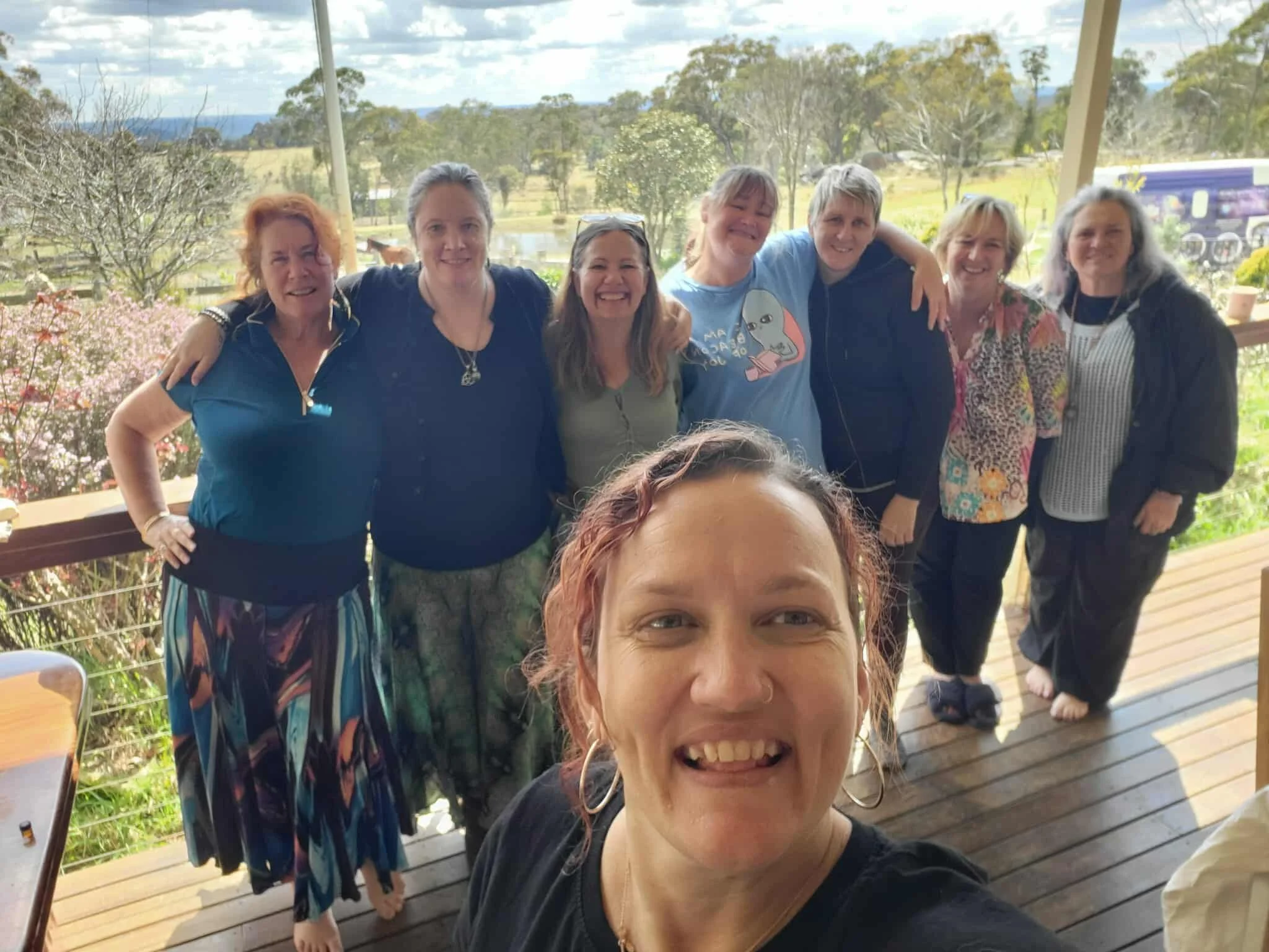 At a retreat a group of nine women smiling and posing for a photo on a porch with a scenic countryside view in the background, loving and supporting themselves and each other being intune and grounded.