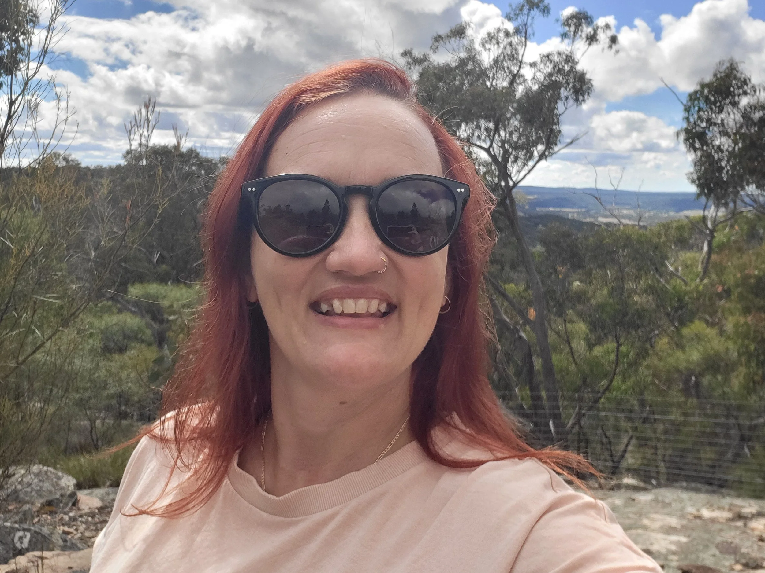 A woman with red hair wearing black sunglasses and a light-colored shirt, smiling outdoors, grounding and reconnecting with herself amongst the trees and a scenic landscape under a cloudy sky in the background.