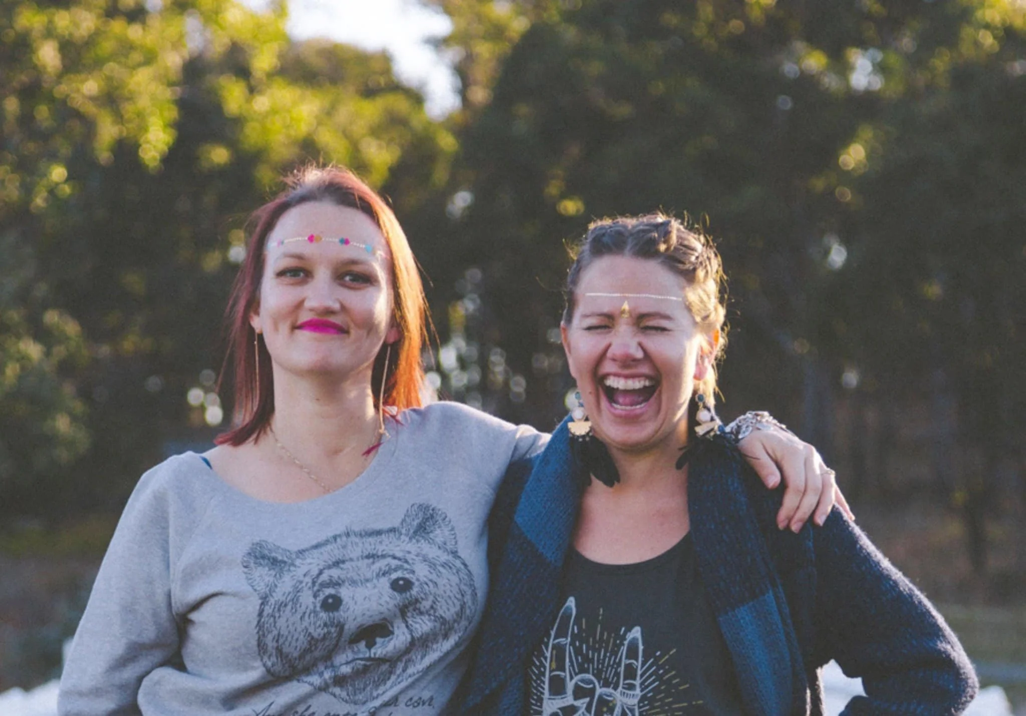 Two women standing outdoors with trees in the background, smiling and having a good time loving and supporting each other, one of them wearing colorful jewelry and the other with large earrings.