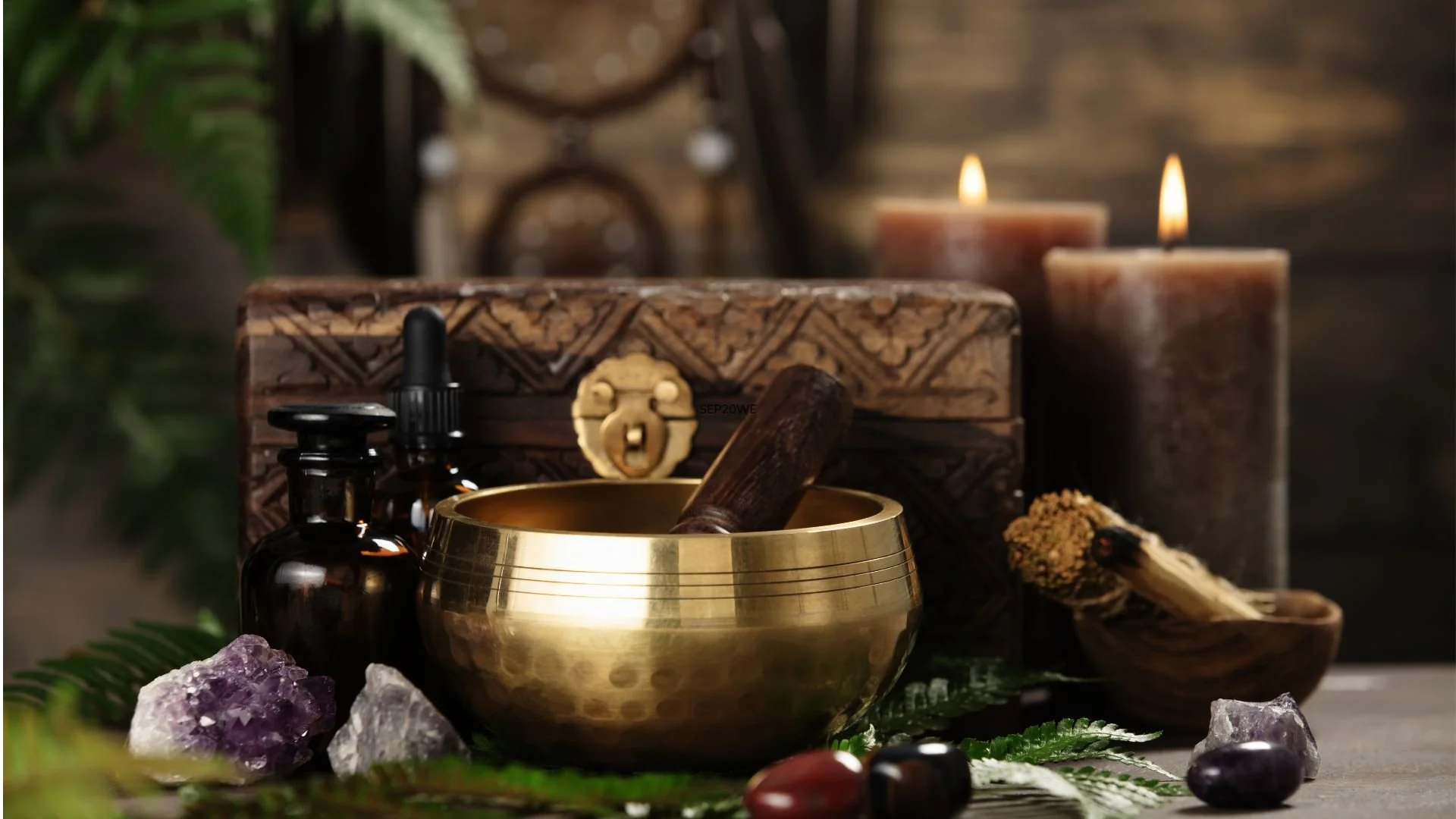 A brass singing bowl with a wooden striker inside, surrounded by purple amethyst and clear quartz crystals, amber essential oil bottles, a carved wooden box, and lit candles in a dimly lit room with greenery.