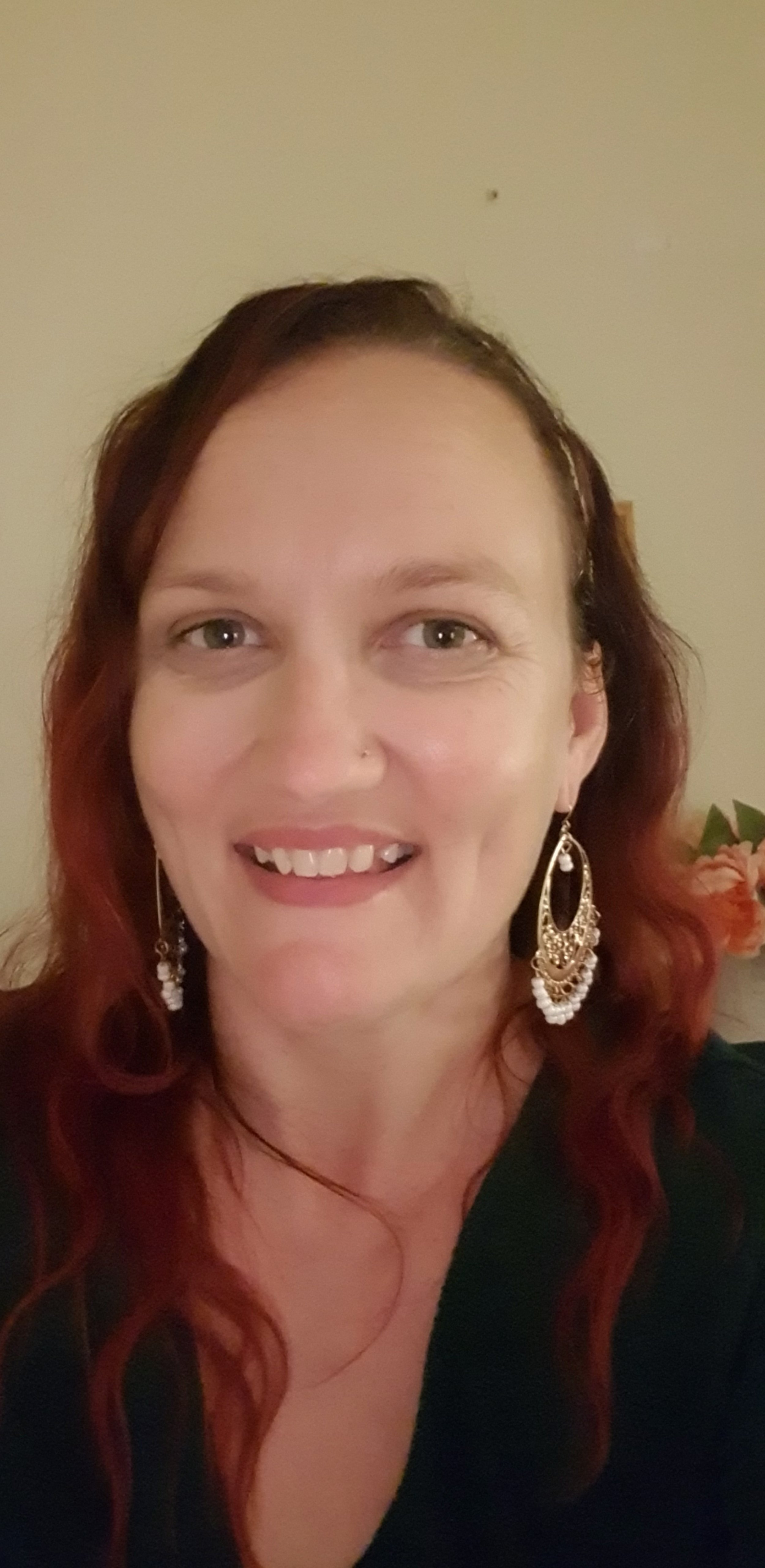 A woman who is a vibrational kinesiologist with light skin, long red hair, and green eyes smiling at the camera. She is wearing large ornate earrings and a black top, with a background that includes a beige wall finally allowing herself to be seen.