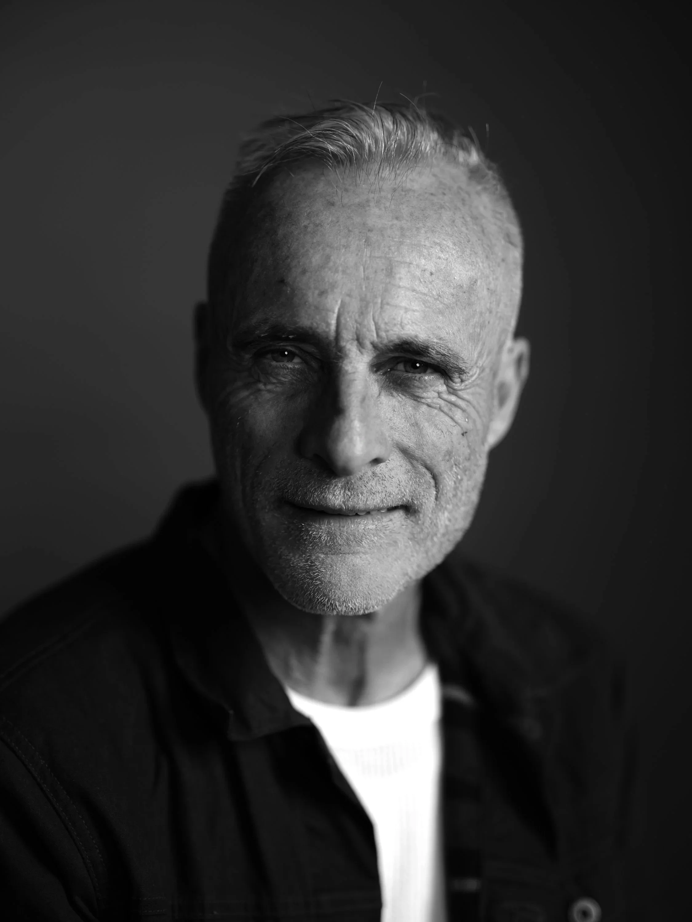 Close-up black and white portrait of a smiling Timothy V. Murphy with short, gray hair, wearing a dark jacket and a white shirt, with a plain dark background.