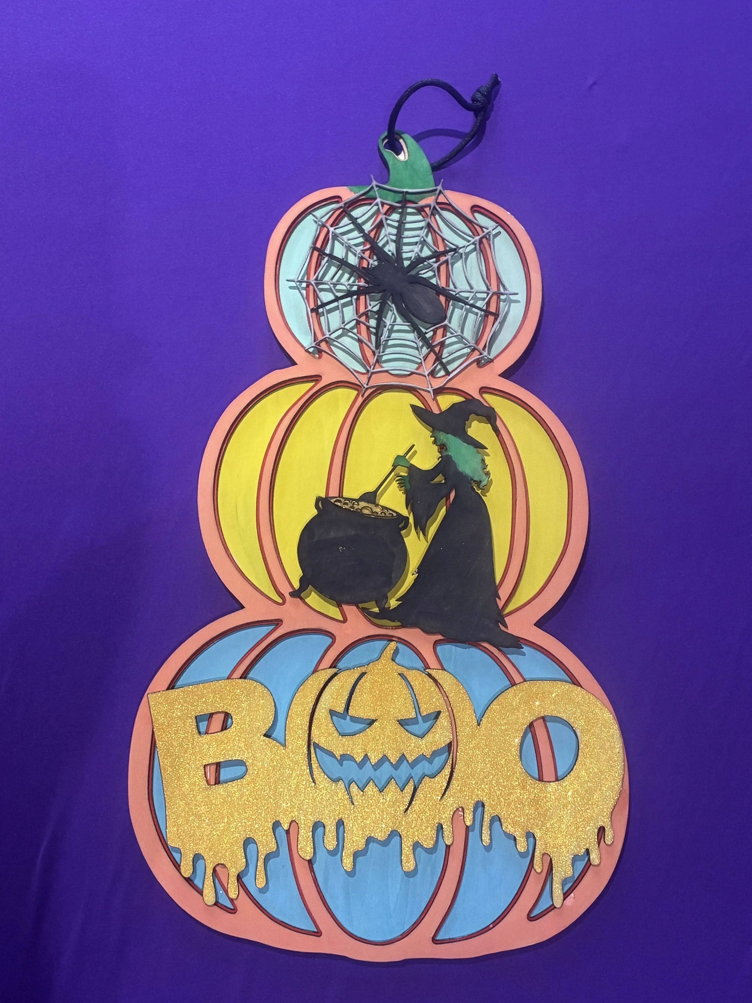 Colorful Halloween-themed decoration with stacked pumpkin-shaped cutouts featuring a witch silhouette, a spider with a web, and a glittery 'Boo' sign.