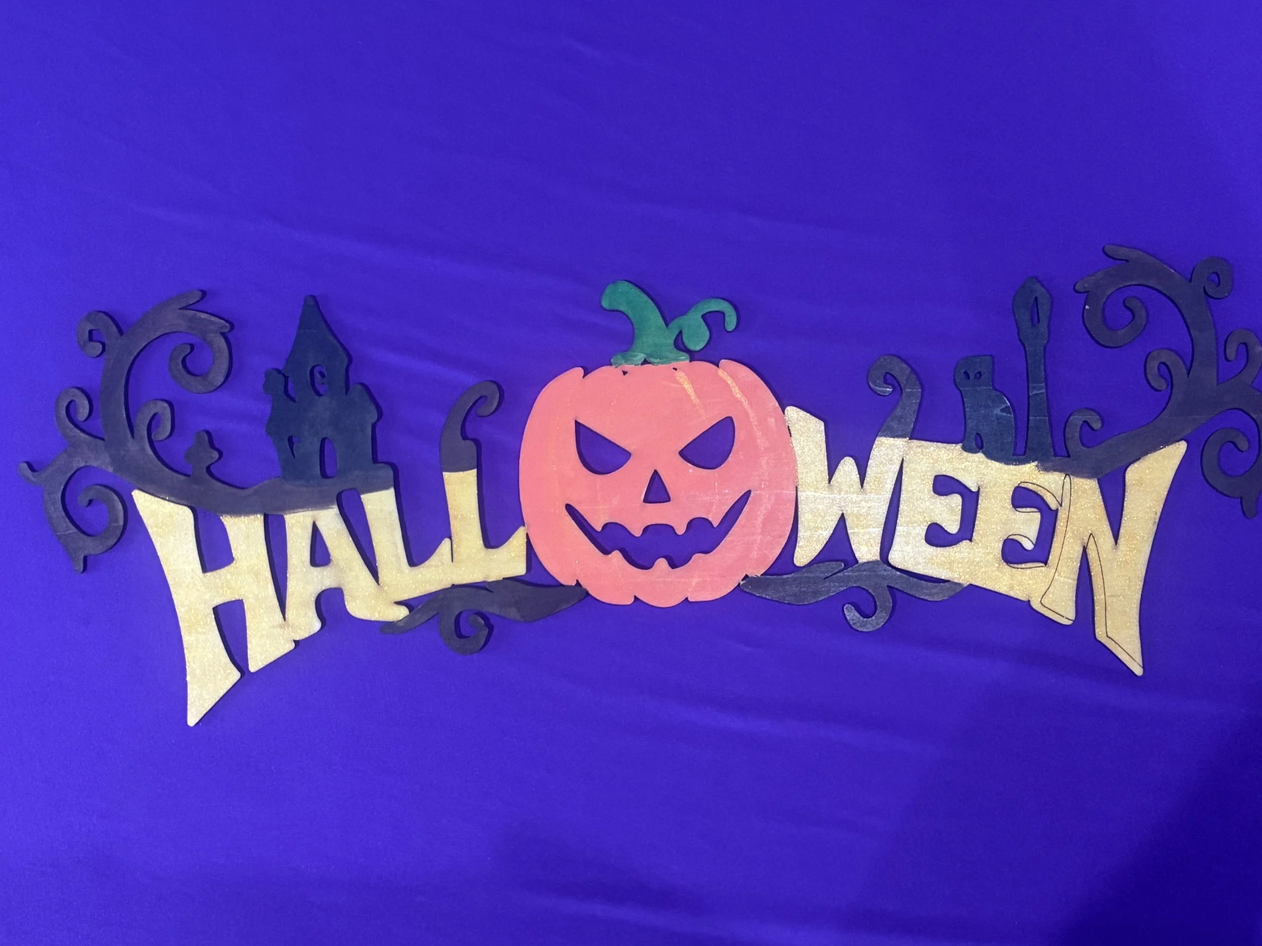 Purple backdrop with black, orange, and gold Halloween-themed decorations, including a smiling pumpkin with a green stem and a wide grin, with cutouts of a haunted house, witches' hats, and swirling patterns.