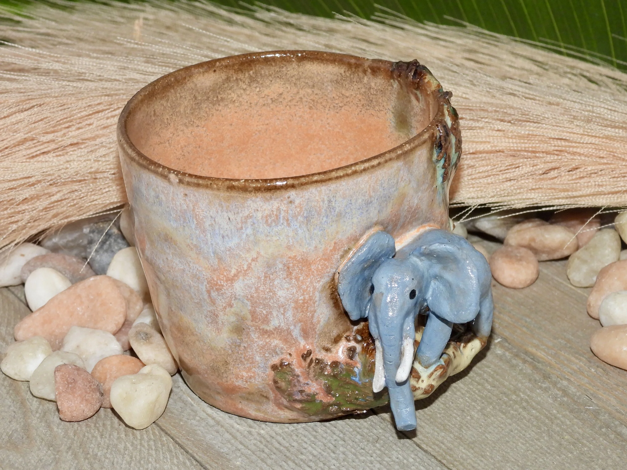 Ceramic mug with an elephant figurine attached to its side, surrounded by small pebbles on a wooden surface.