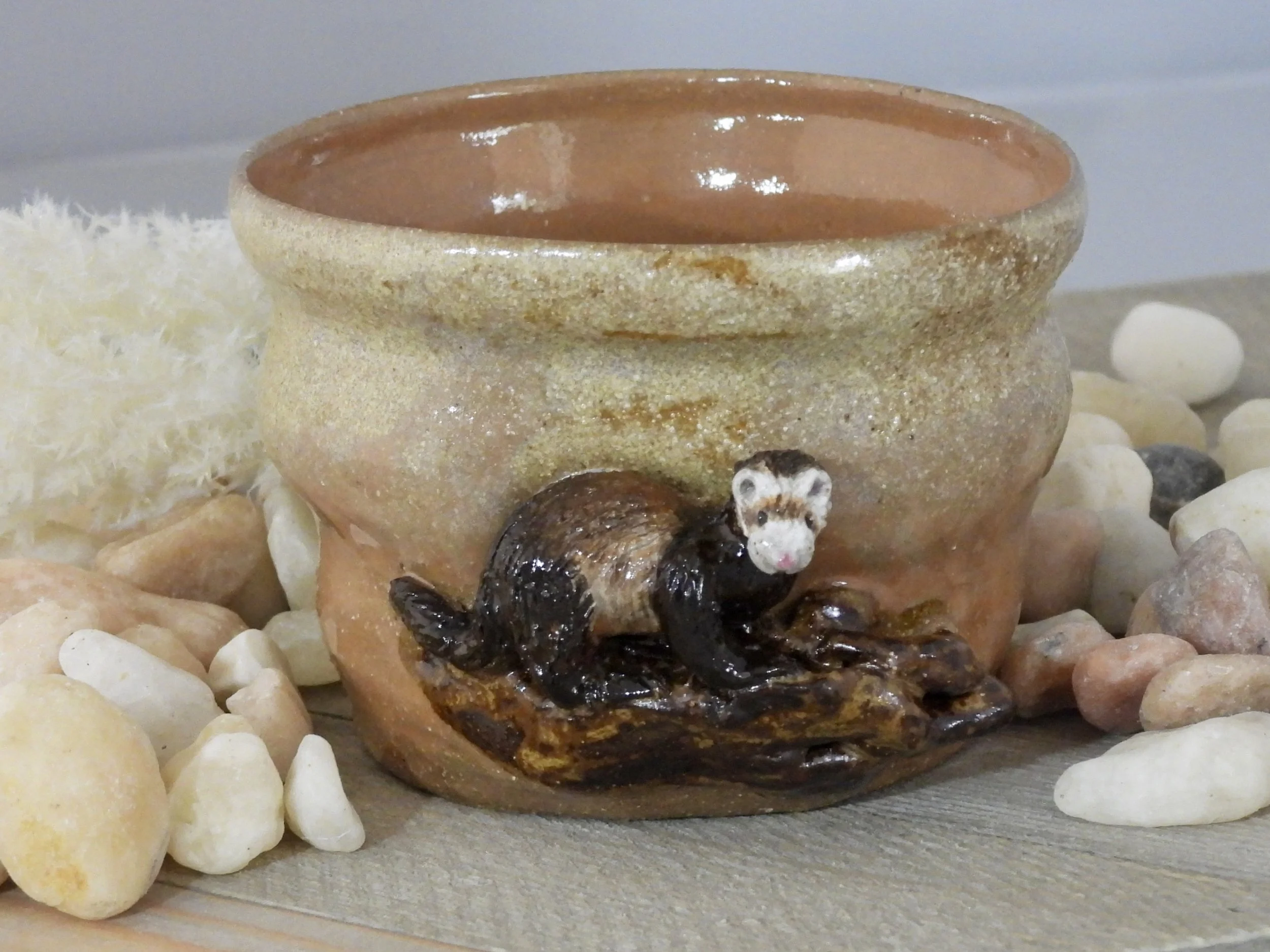 A small decorative ceramic bowl with a bear figurine attached to its base, surrounded by various small white and tan stones.