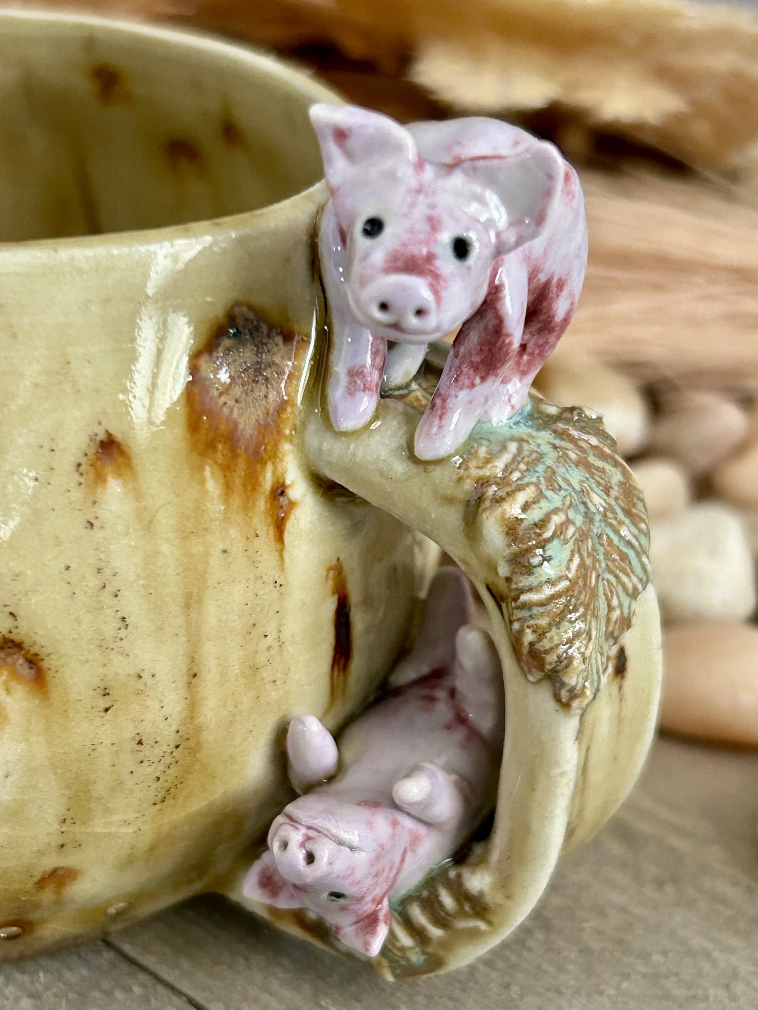 Ceramic mug with pig-shaped decorations, featuring a pig's head and paws emerging from the mug, with a rustic wooden surface in the background.