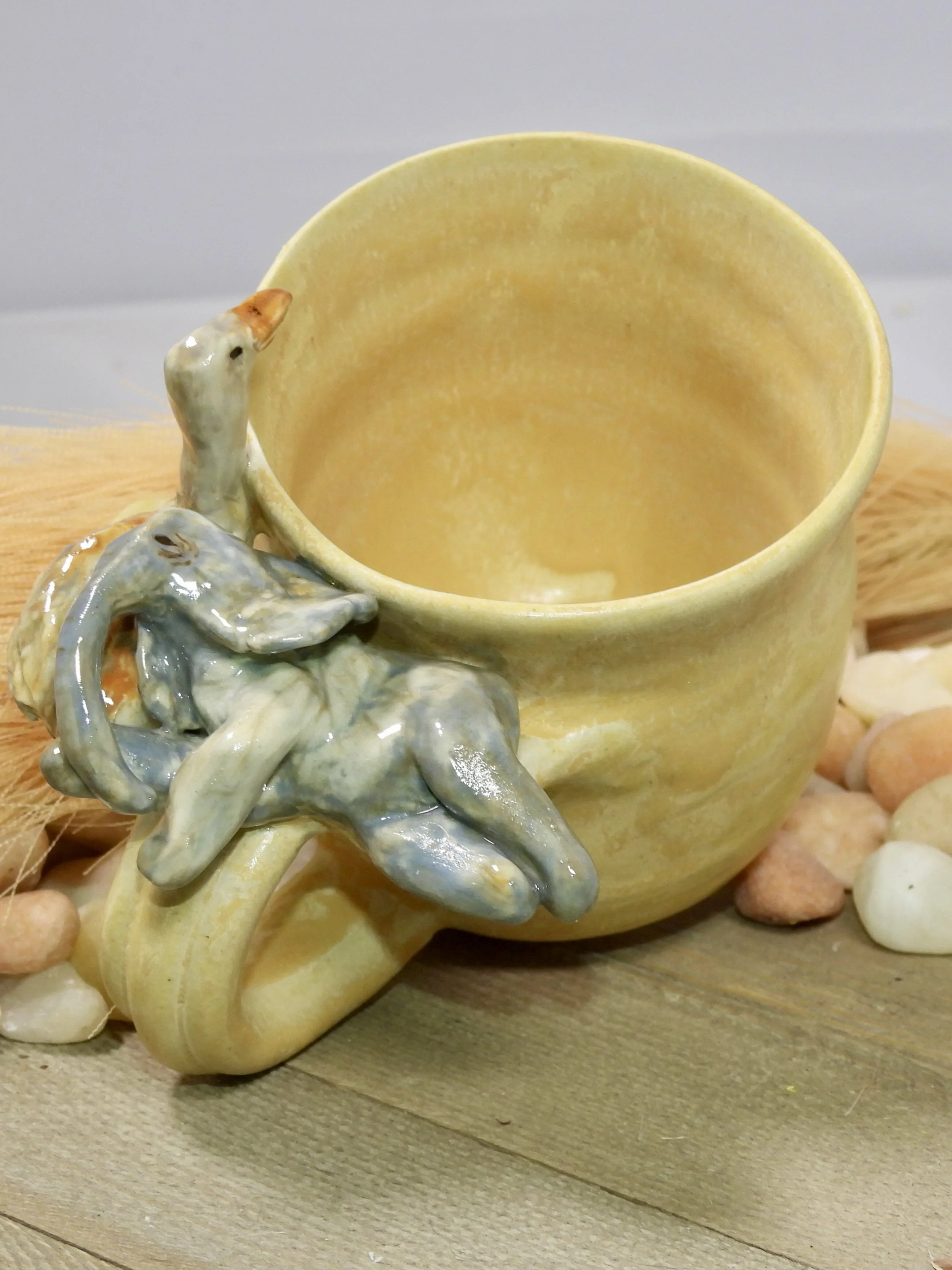 Ceramic mug with a decorative baby duck sculpture attached to its handle, placed on a wooden surface surrounded by small rocks.