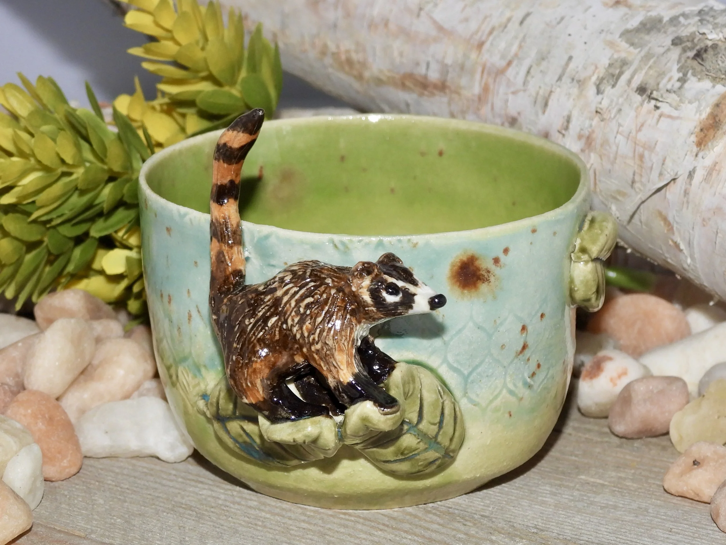 A ceramic cup with a painted last and a 3D meerkat figure sitting on a branch attached to the cup. The cup is light green on the inside and pale green on the outside, surrounded by decorative rocks and green foliage.