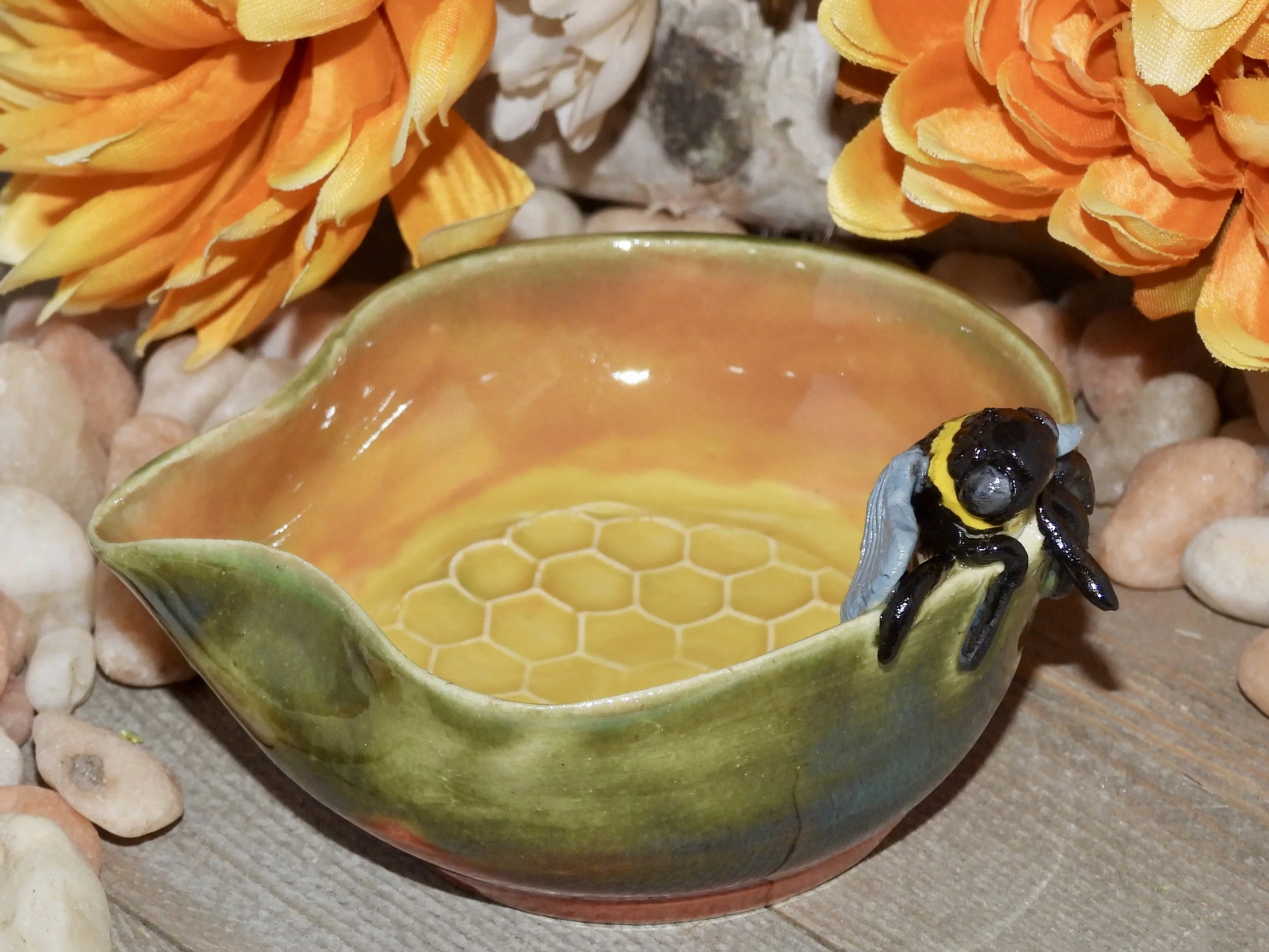 A ceramic bowl with a honeycomb design inside, decorated to resemble a flower, with a bee figurine on its rim, surrounded by artificial orange flowers and small pebbles.