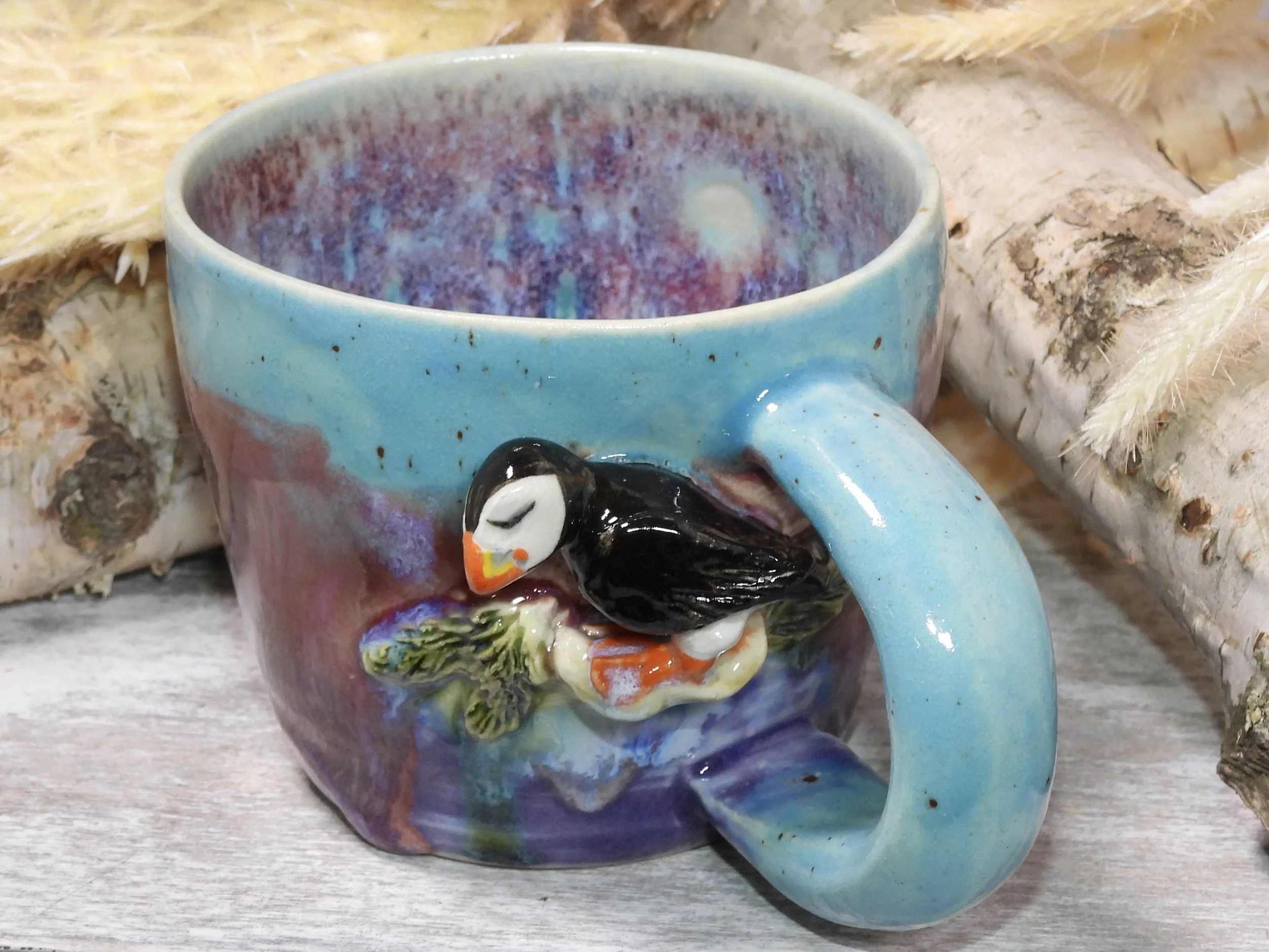 Colorful ceramic mug with a 3D duck decoration on its side, sitting on a wooden surface with logs in the background.