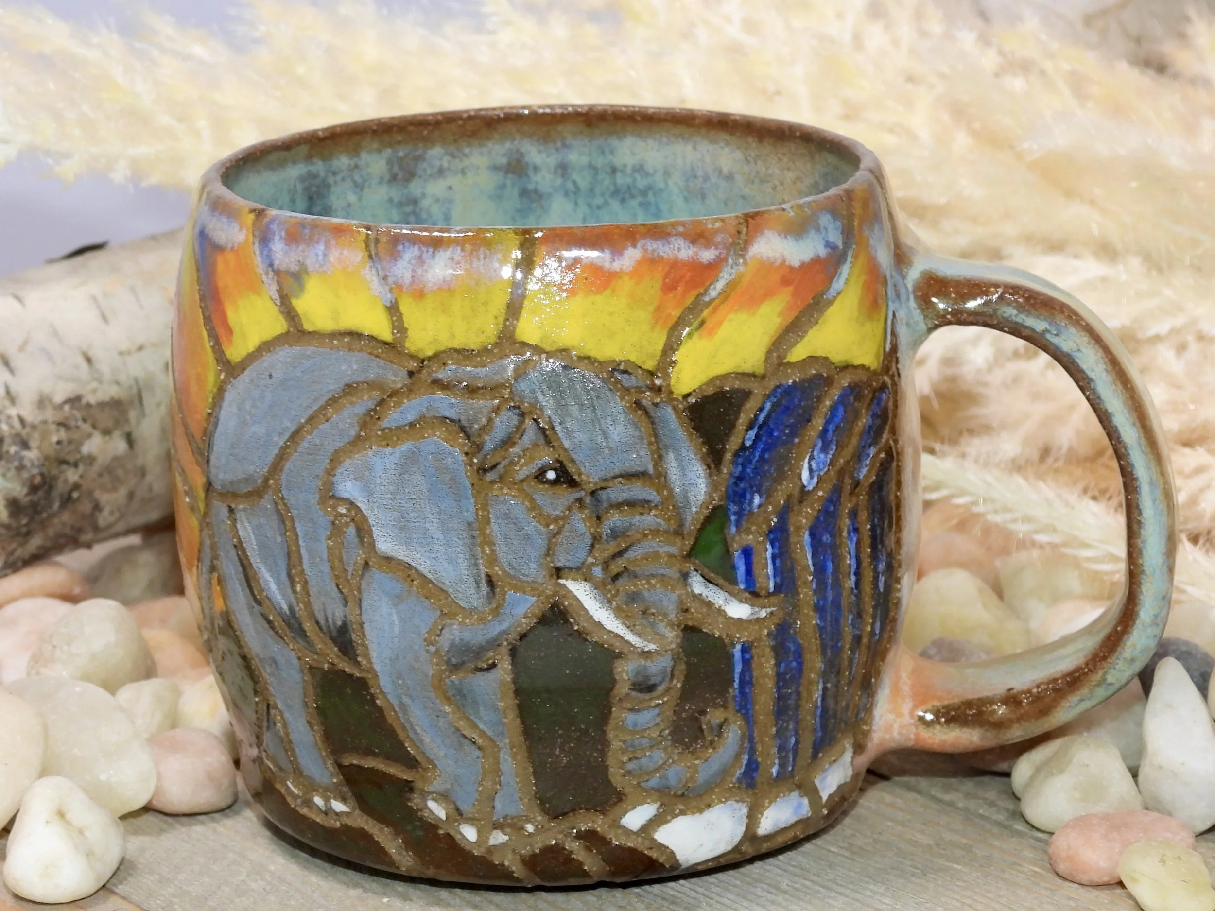 A ceramic mug decorated with an elephant design and a colorful sunset in the background, set on a wooden surface surrounded by small pebbles.