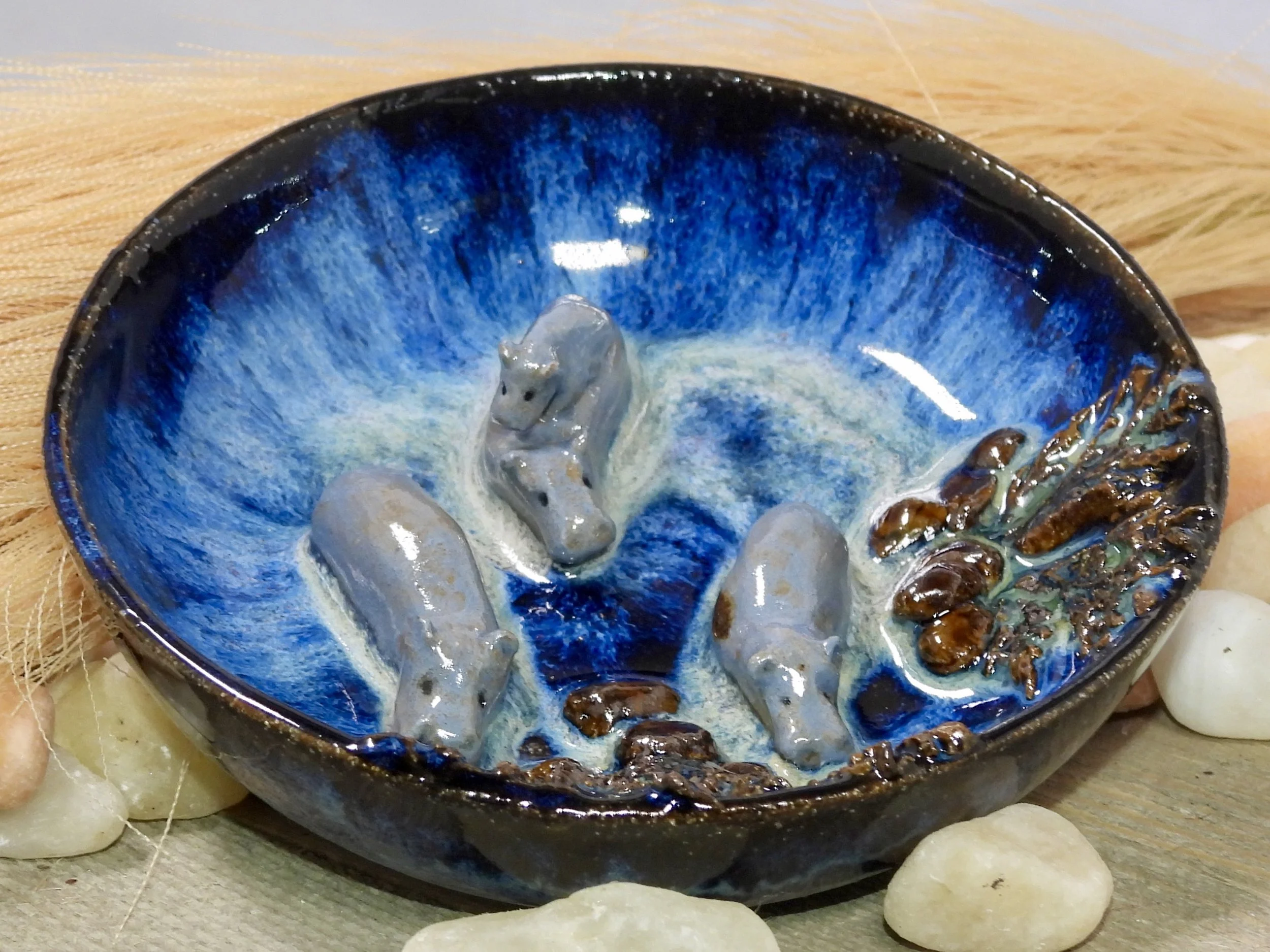 A ceramic bowl with a blue and white glaze design, containing three small irregularly shaped stones or stones-like objects, with a cluster of small dried plants on the side, placed on a wooden surface with other scattered stones around.
