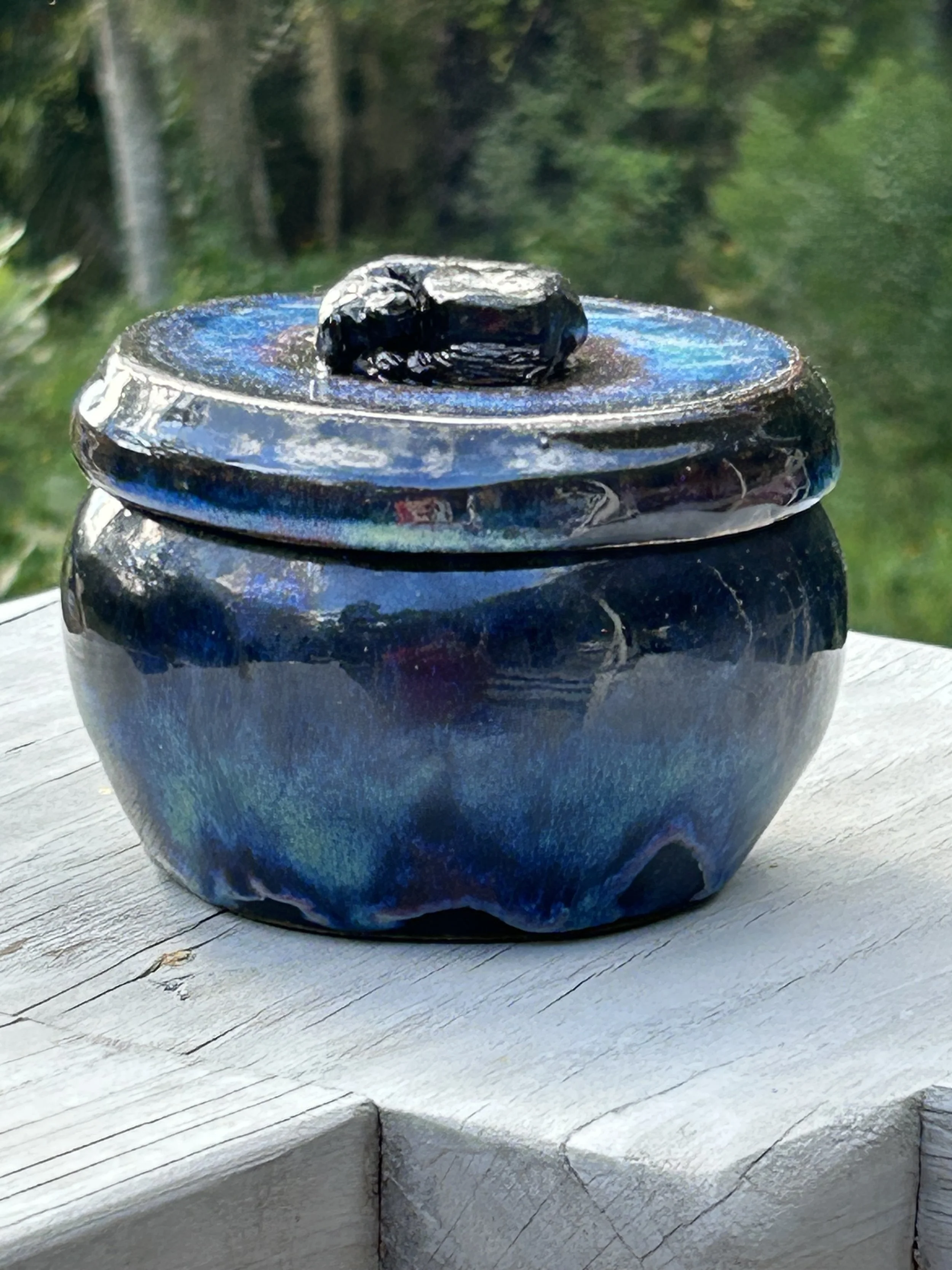 A decorative jar with a shiny, iridescent blue and black surface, topped with a closed lid and a black stone on top, placed on a white wooden surface outdoors.
