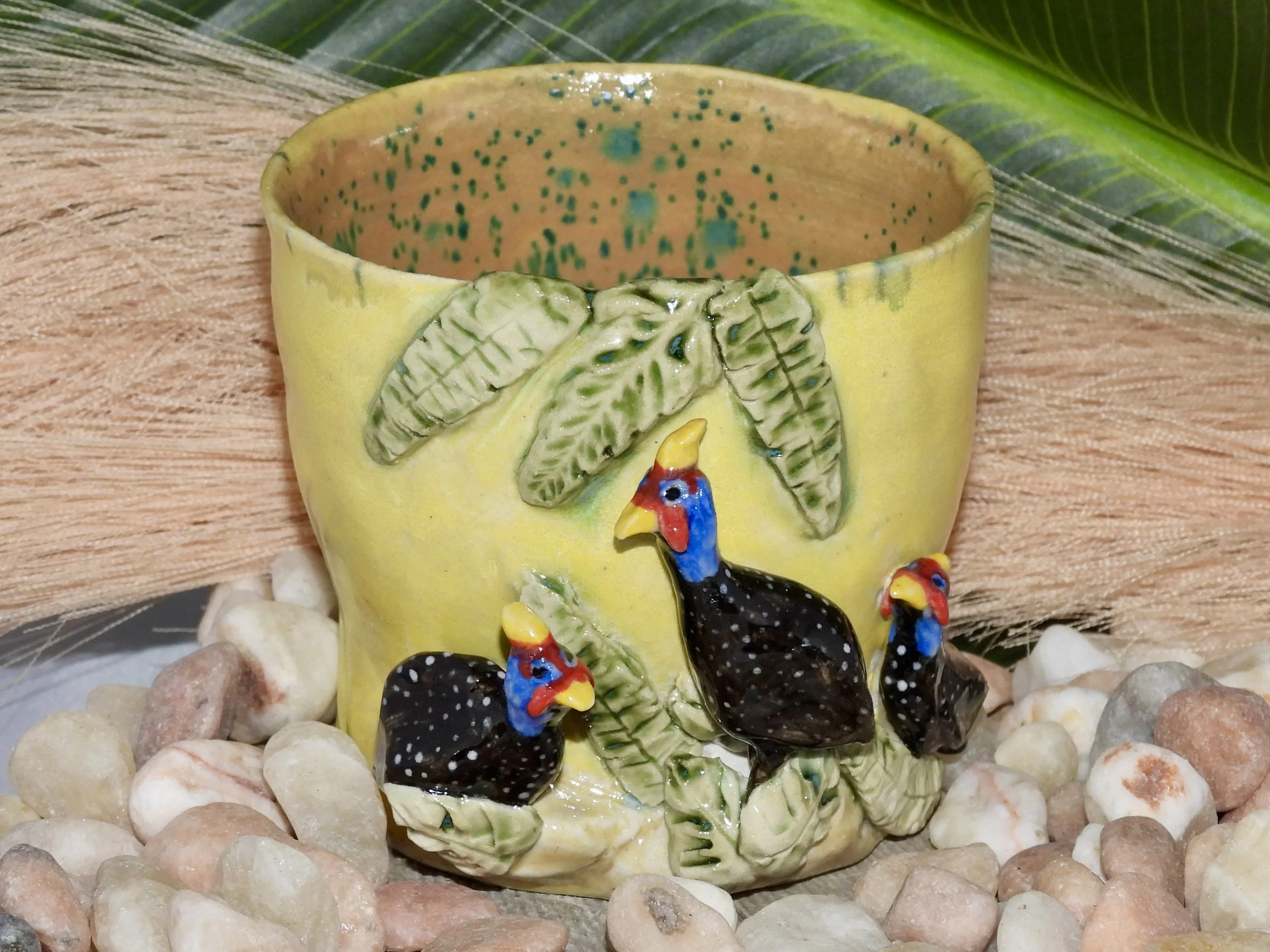 Decorative ceramic mug with a yellow background and three black roosters with red and blue heads, adorned with green leaves, surrounded by small rocks.