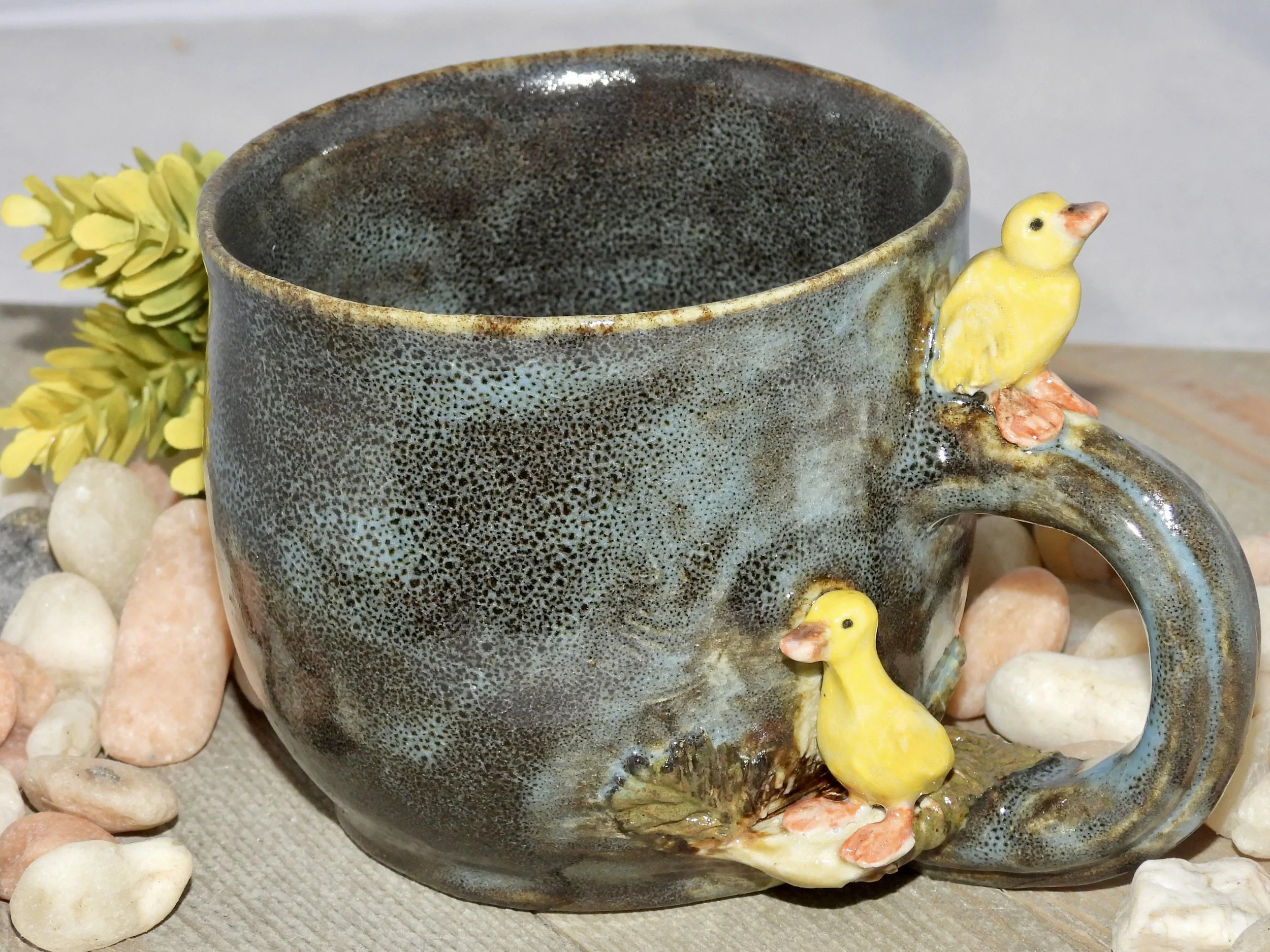 A decorative ceramic mug with a textured, dark glaze surface. It features two small, yellow ceramic ducklings attached to the handle and one duckling on the base of the handle. The mug is placed on a surface surrounded by small, colorful stones and a