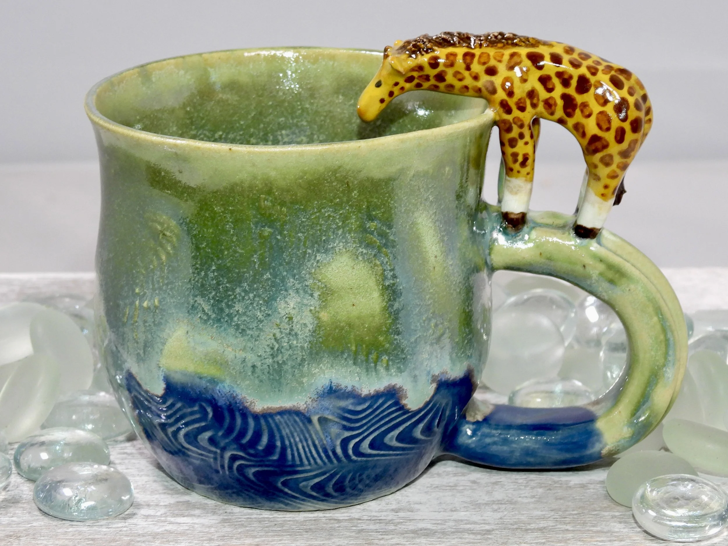 A ceramic mug with a green and blue ocean wave design, featuring a 3D giraffe figurine on the handle, surrounded by glass pebbles.