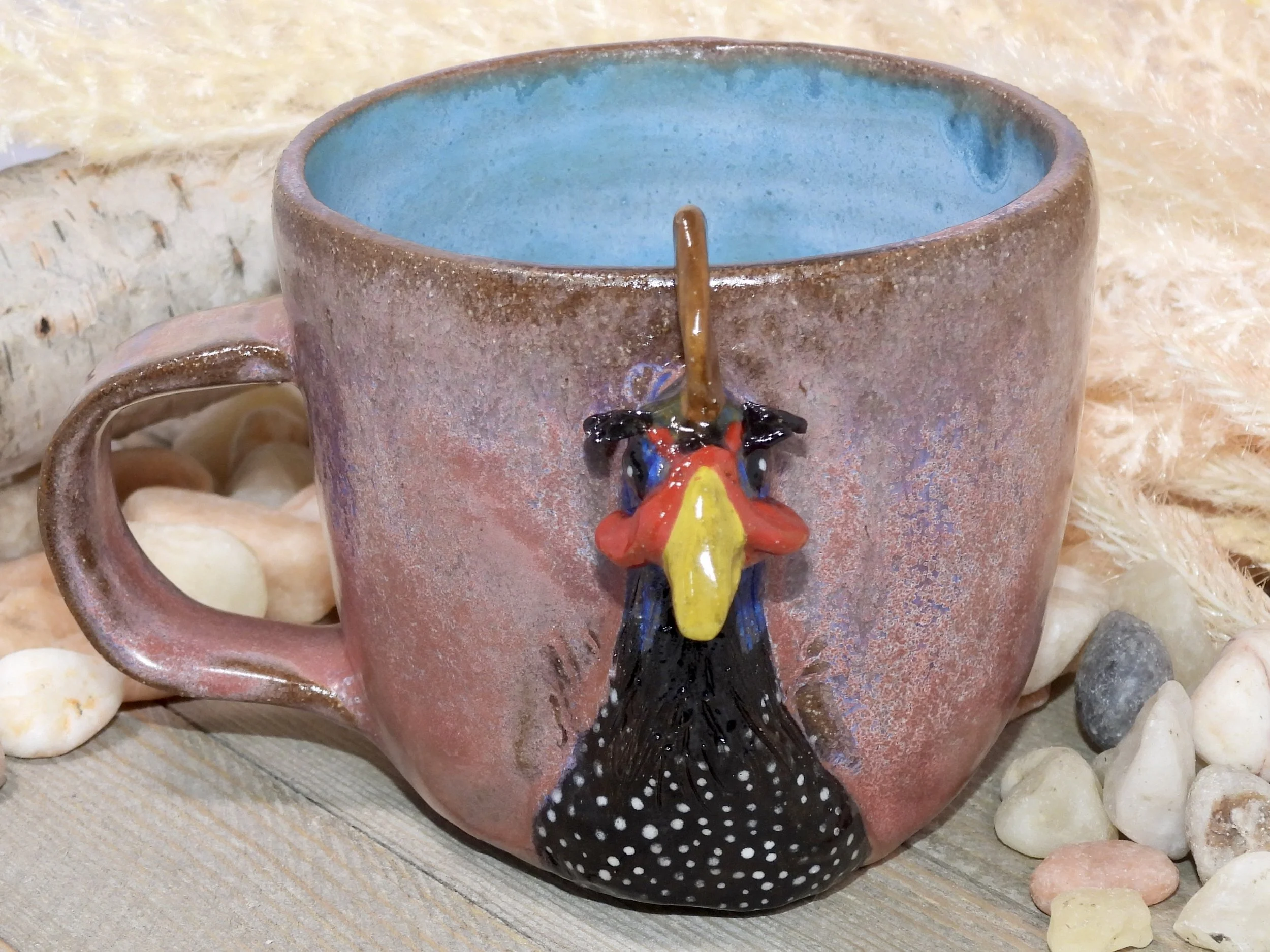 A ceramic mug with a rooster head handle, pinkish body, blue interior, surrounded by rocks on a wooden surface.