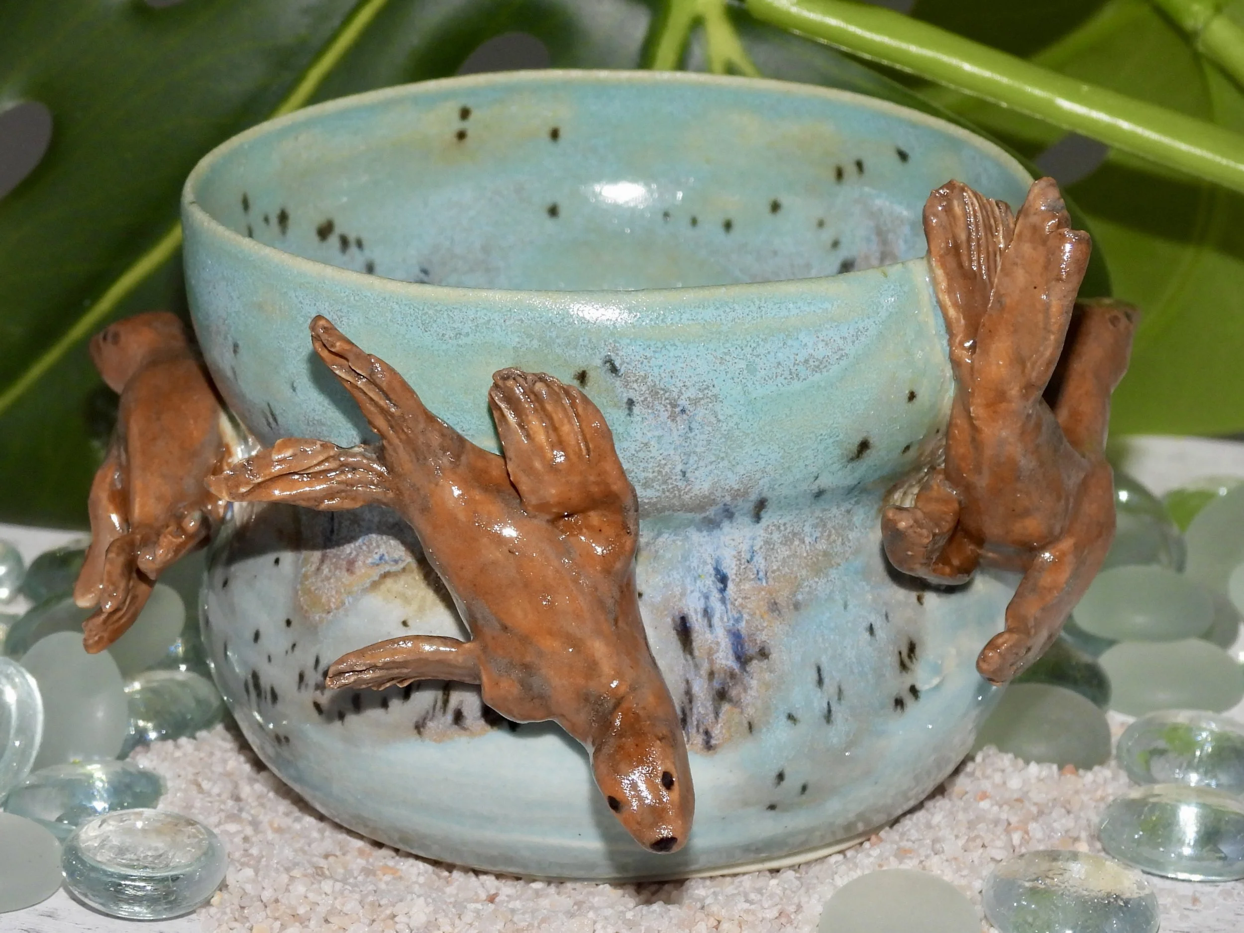 A ceramic bowl with a glaze in shades of blue and black spots, decorated with three small, detailed brown hands sculpted around it, resting on a surface with round glass pebbles and a leafy background.