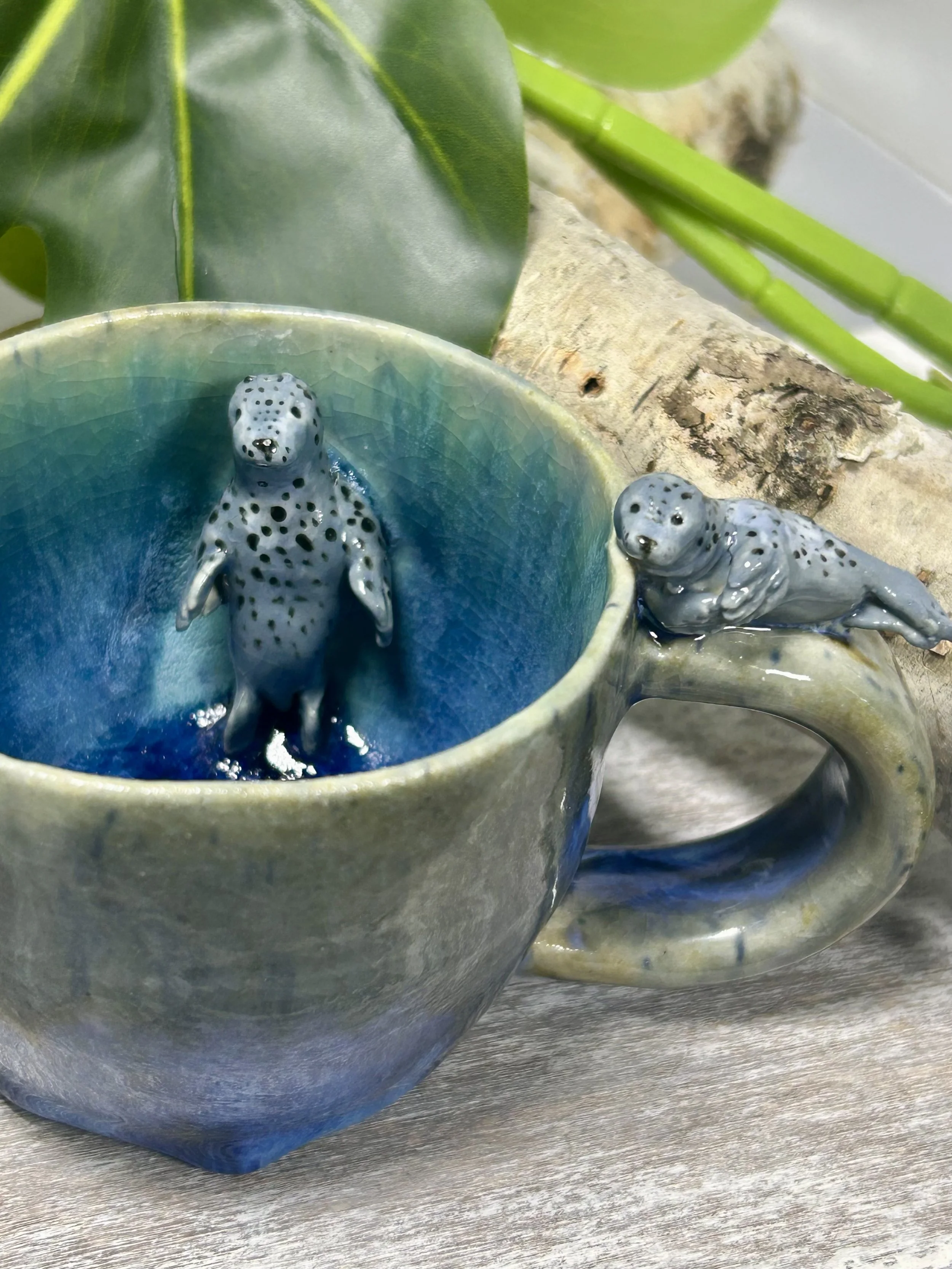 A ceramic mug with a blue and green glaze features two small ceramic animal figurines resembling spotted seals or otters, one inside the mug and one hanging over the mug's handle, set on a wooden surface with green foliage in the background.
