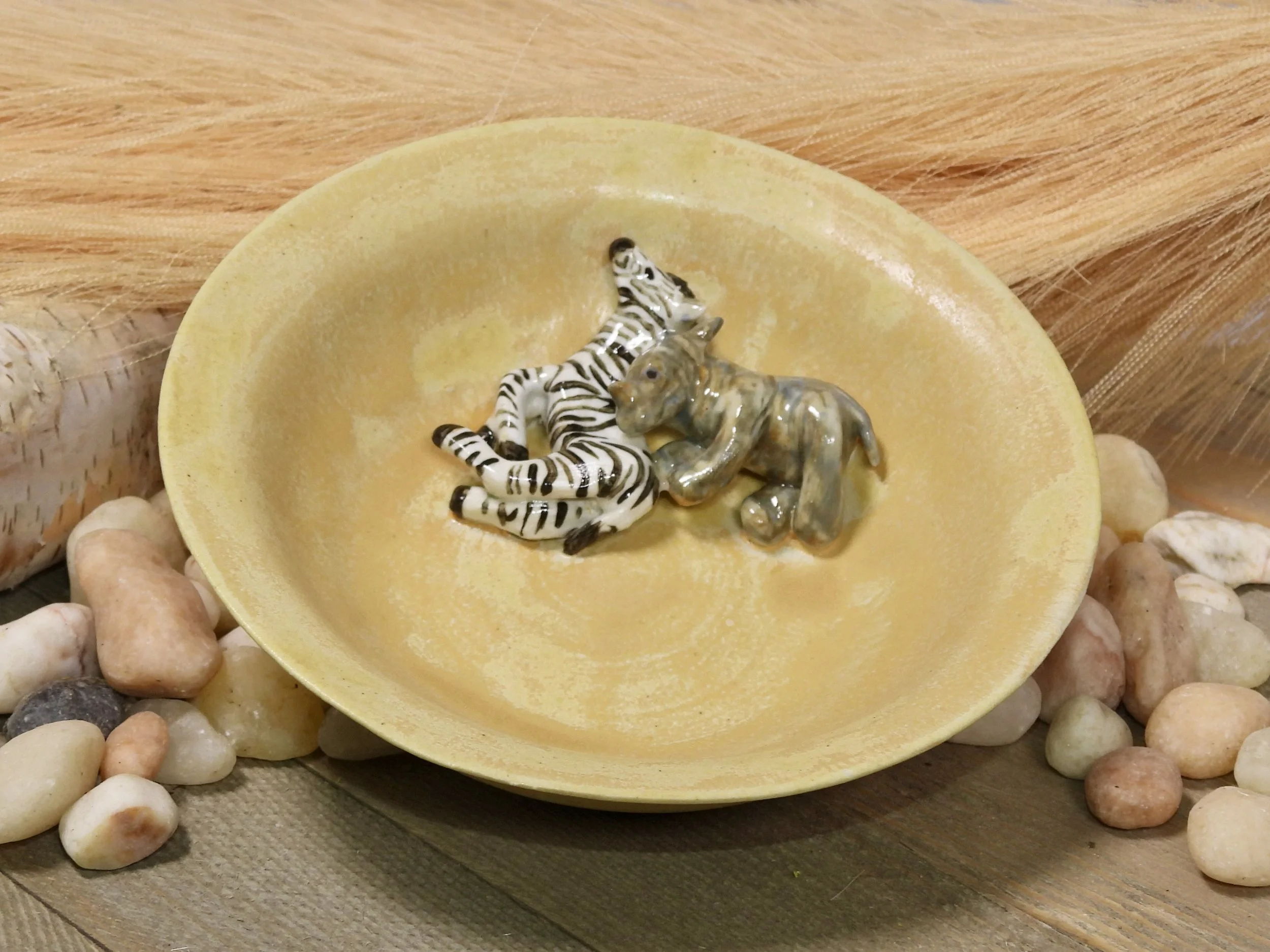 Decorative yellow bowl with a small zebra figurine and a small elephant figurine inside, surrounded by smooth stones and wooden surface.